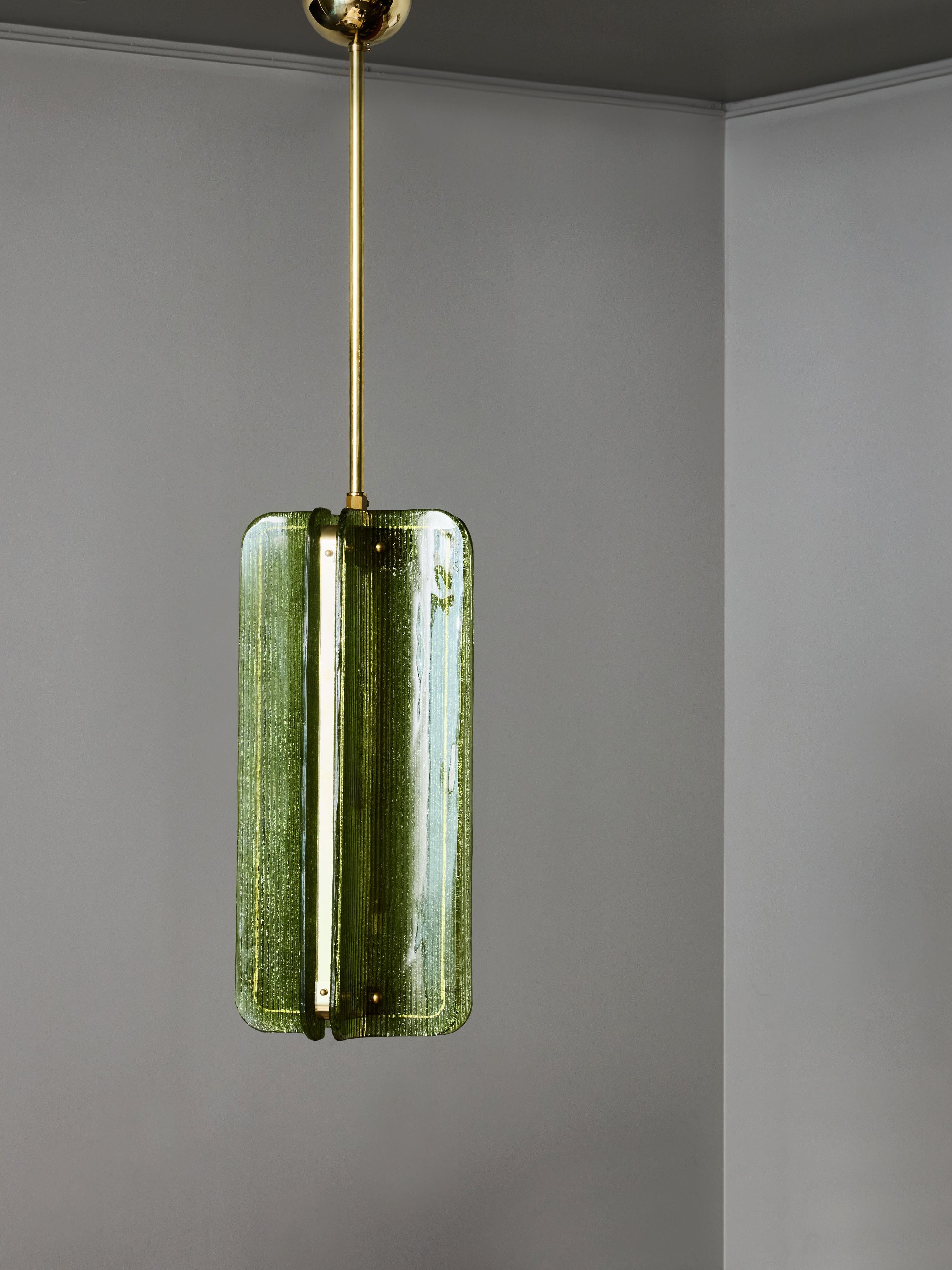 Una sofisticada lámpara colgante moderna, con un conjunto escultural de paneles de cristal texturizado montados alrededor de una estructura central de latón. La lámpara se compone de cuatro elementos de cristal curvados verticalmente, cada uno con