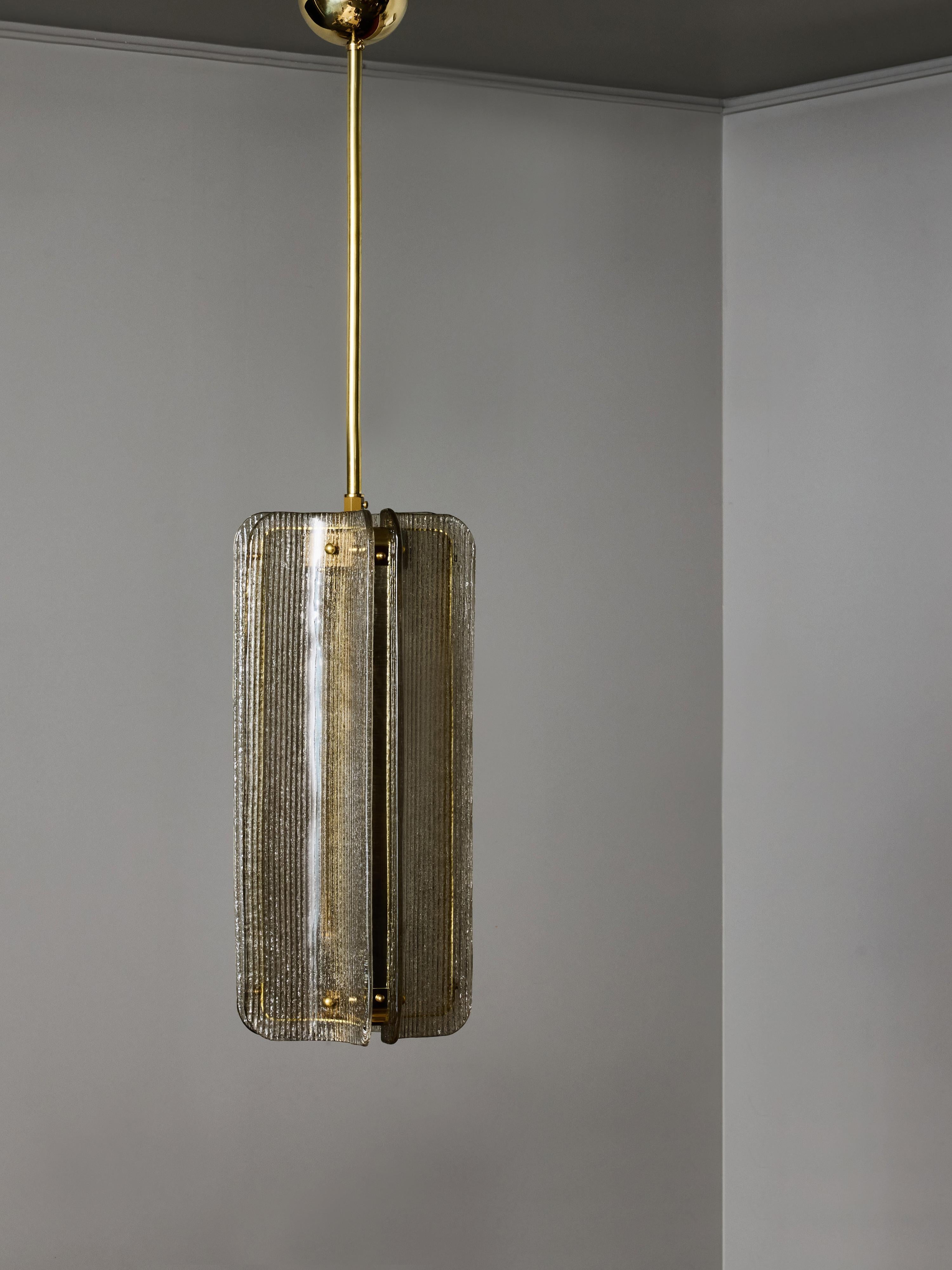 Lampe suspendue moderne et sophistiquée, composée d'un assemblage sculptural de panneaux de verre texturé montés autour d'une structure centrale en laiton. La lampe est composée de quatre éléments en verre incurvés verticalement, chacun présentant