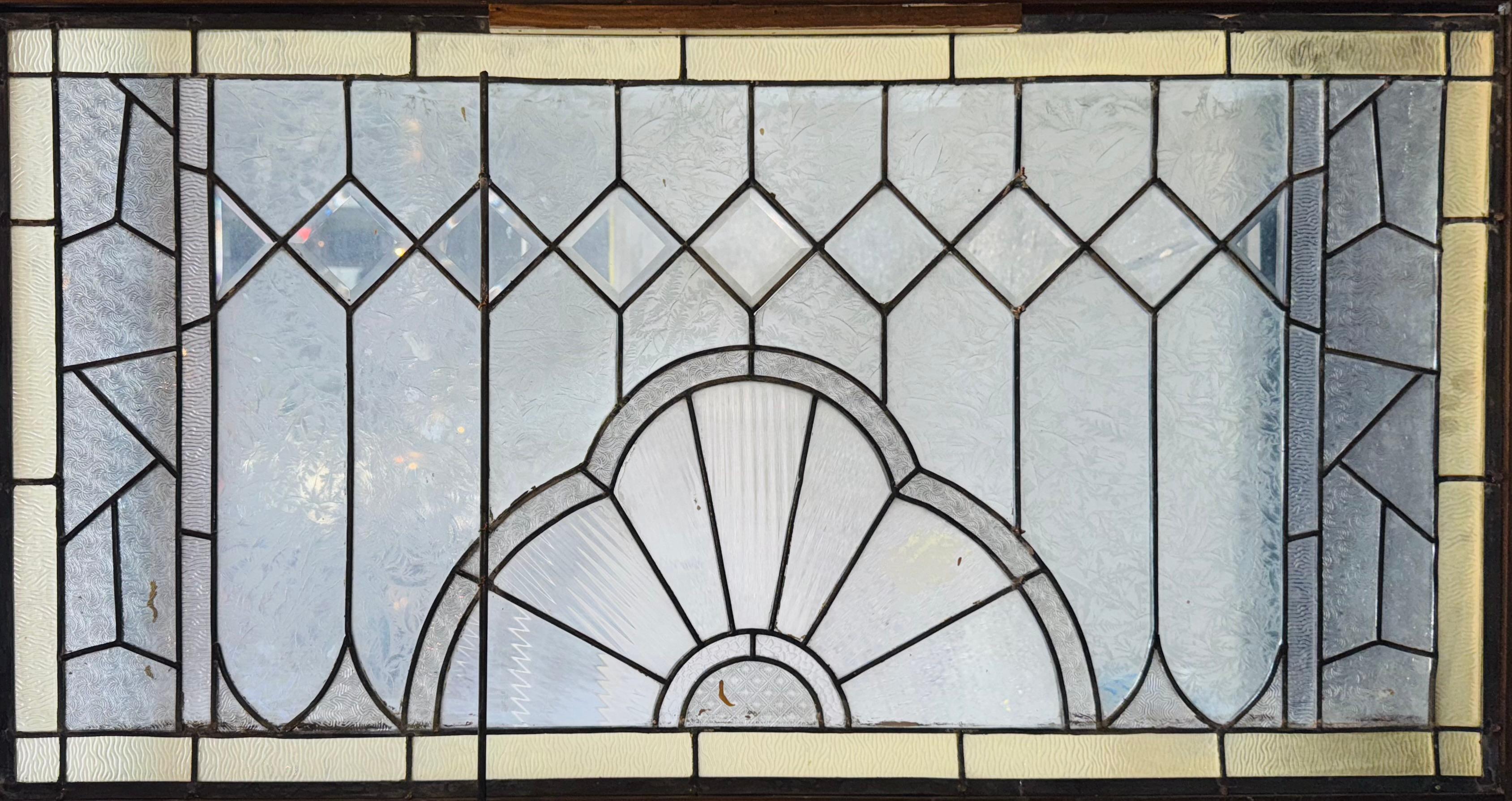 Fenêtre d'imposte de quatre pieds de long avec verre clair texturé et bordure teintée jaune clair. Au centre se trouve un motif en éventail avec un verre biseauté en forme de diamant au-dessus, et des motifs géométriques de part et