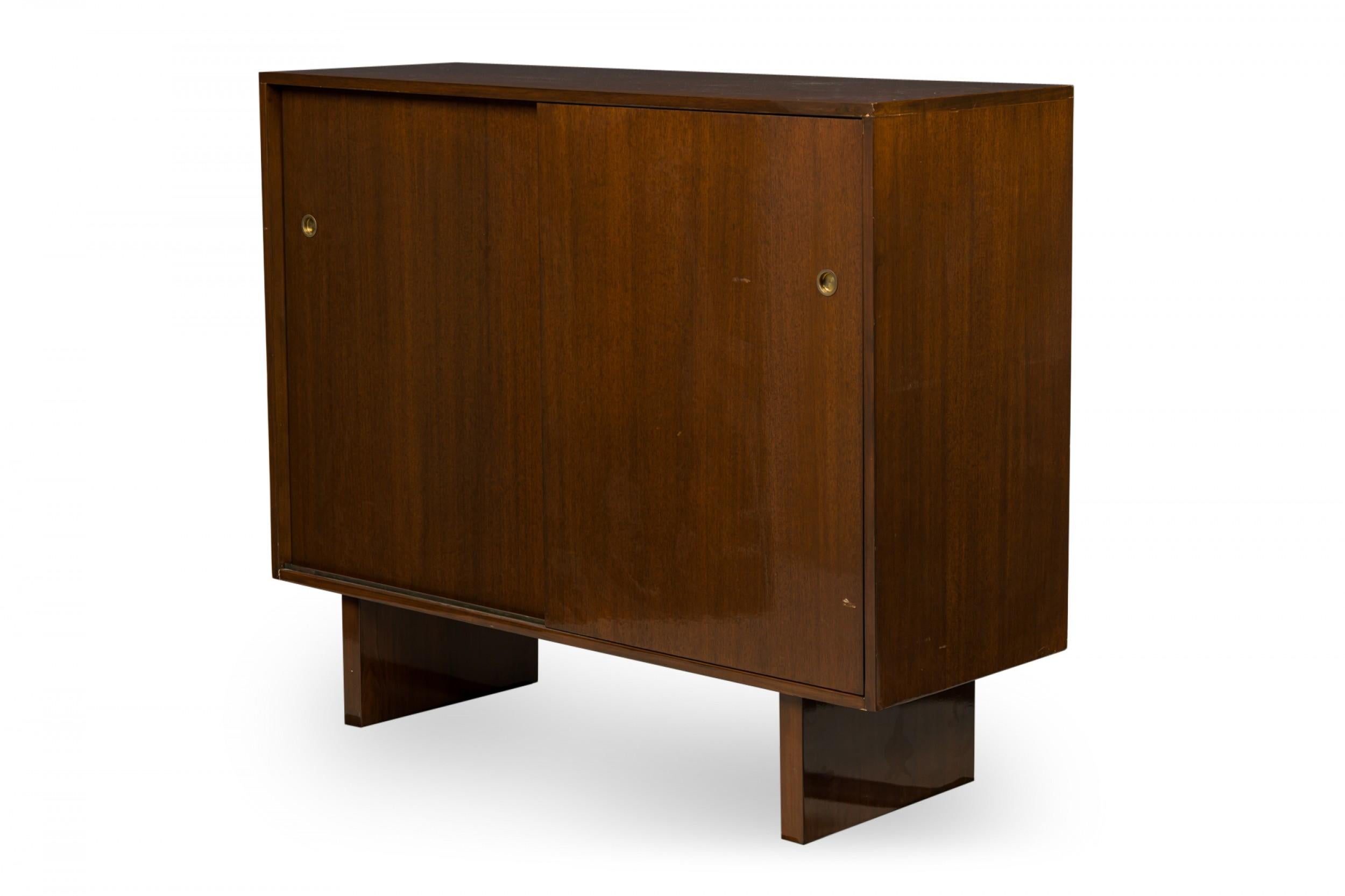 American Mid-Century Modern mahogany cabinet with two sliding doors with inset brass ring door pulls, which slide open to reveal a multi-compartment interior with pull out drawers, resting on two wide plank legs. (T.H. ROBSJOHN-GIBBINGS).
