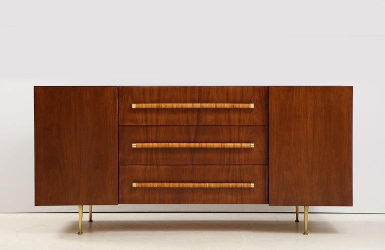 T.H. Robsjohn-Gibbings for Widdicomb rare 1950s sideboard or cabinet in rich deep walnut including central drawers with original circular woven rattan handles and brass end caps, cabinets on each end with adjustable shelves, and brass legs. This