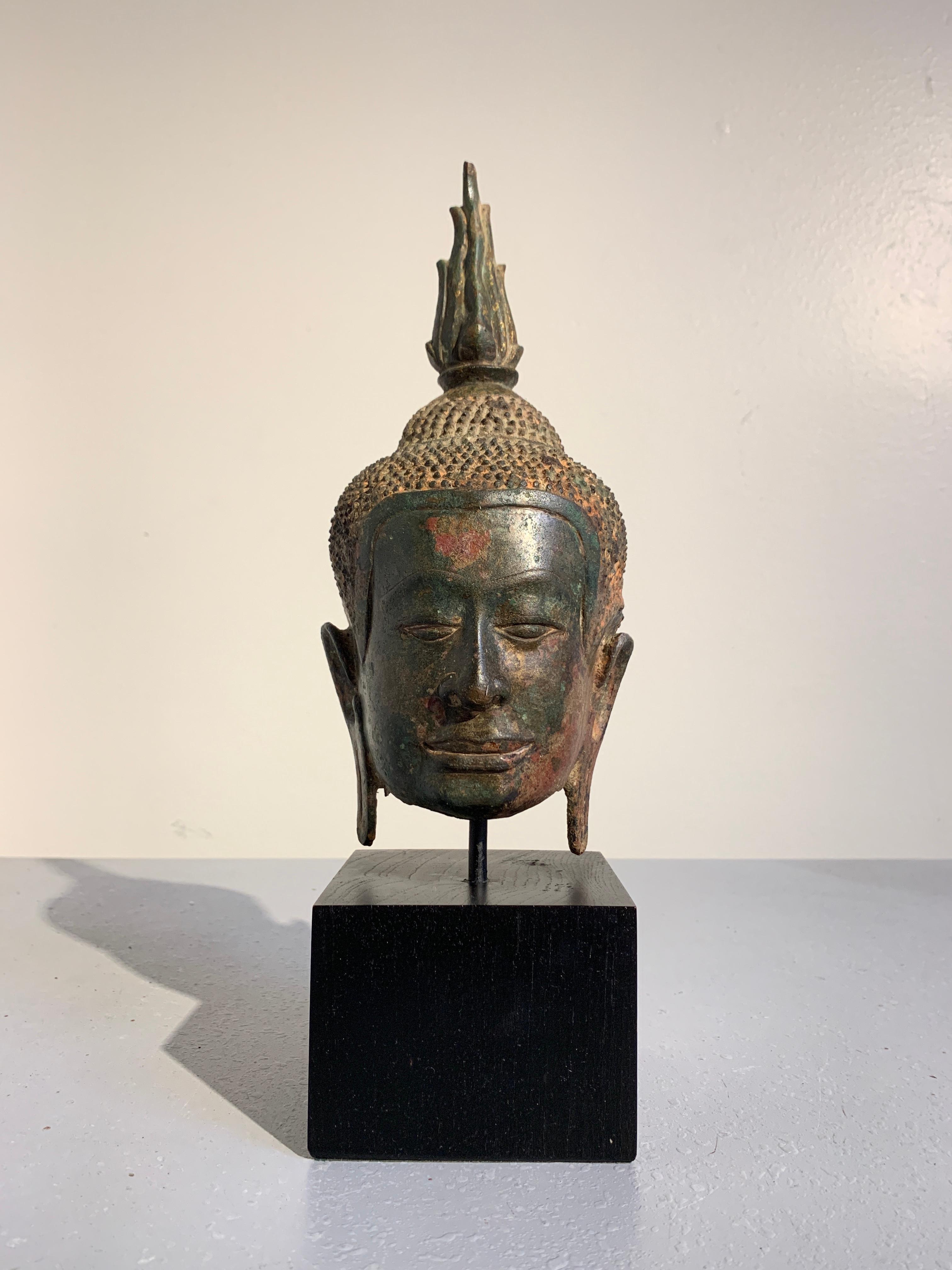 A beautifully patinated bronze Buddha head from the Kingdom of Ayutthaya, Thailand, circa 14th century.

The Buddha's face has a distinct Khmer influence, with a square face and jaw, broad nose, and long straight lips curled up ever so slightly at