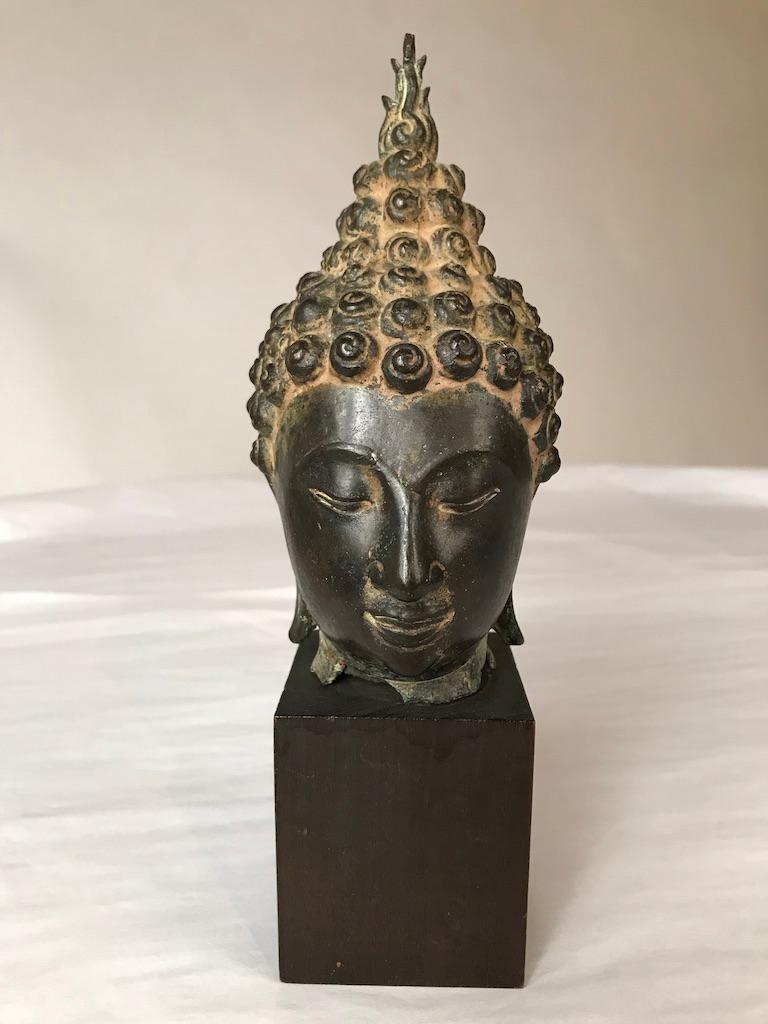 A Thai, Ayutthaya style bronze head of Buddha Shakyamuni, late 16th or early 17th century. With a serene expression on his face, downcast eyes, arched eyebrows, smiling lips, with curled headdress and ushnisha topped by a flame. Measures: 9.25