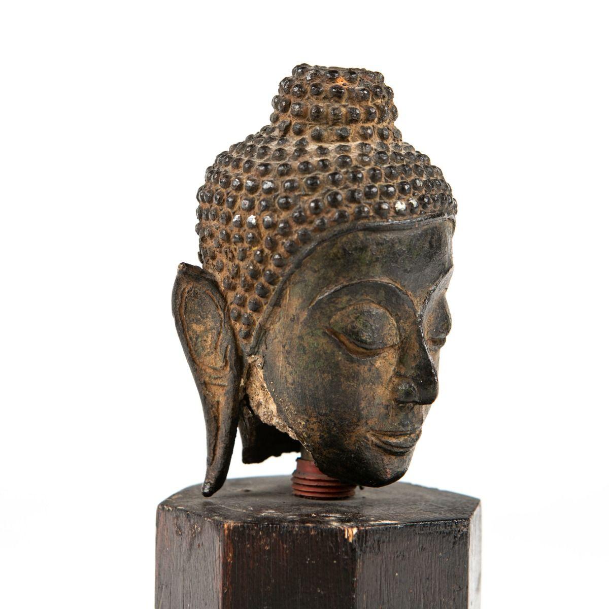 A small Buddha bronze head, measuring 11 cm in height, originating from Thailand (1600-1700 AD).
It is loosely mounted on a hexagonal wooden base.
This elegant and decorative head features a naturally aged patina, giving it an authentic and timeless