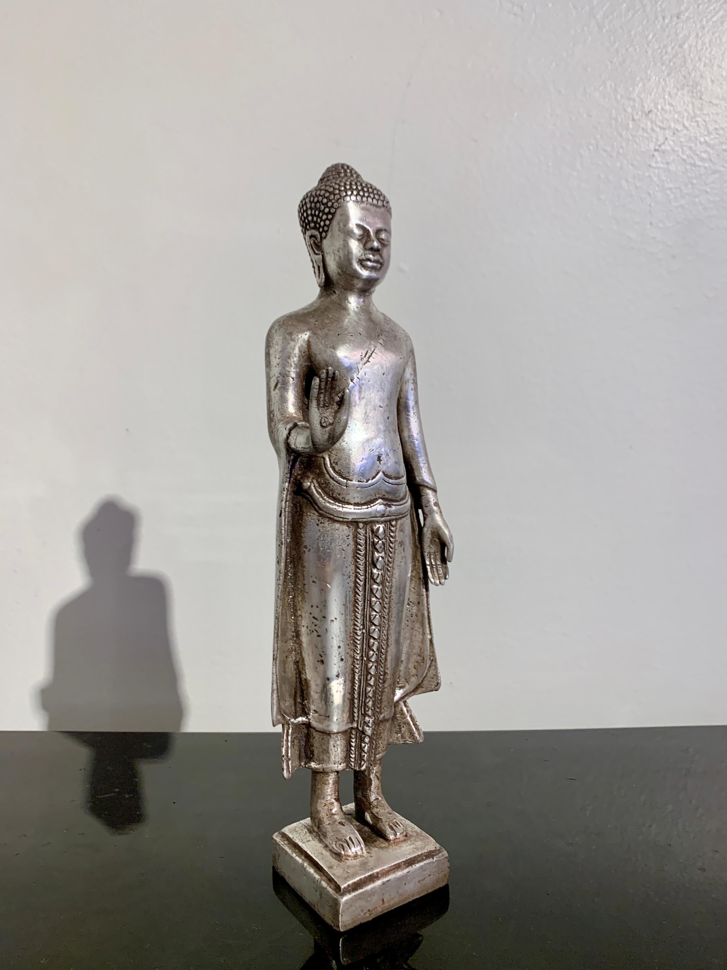 An impressive and gracious Thai cast silver alloy standing Buddha, mid 20th century, Thailand.

The Buddha stands upright upon a small square plinth. He is dressed in layered robes, including a dhoti covering his legs, and a sanghati wrapped