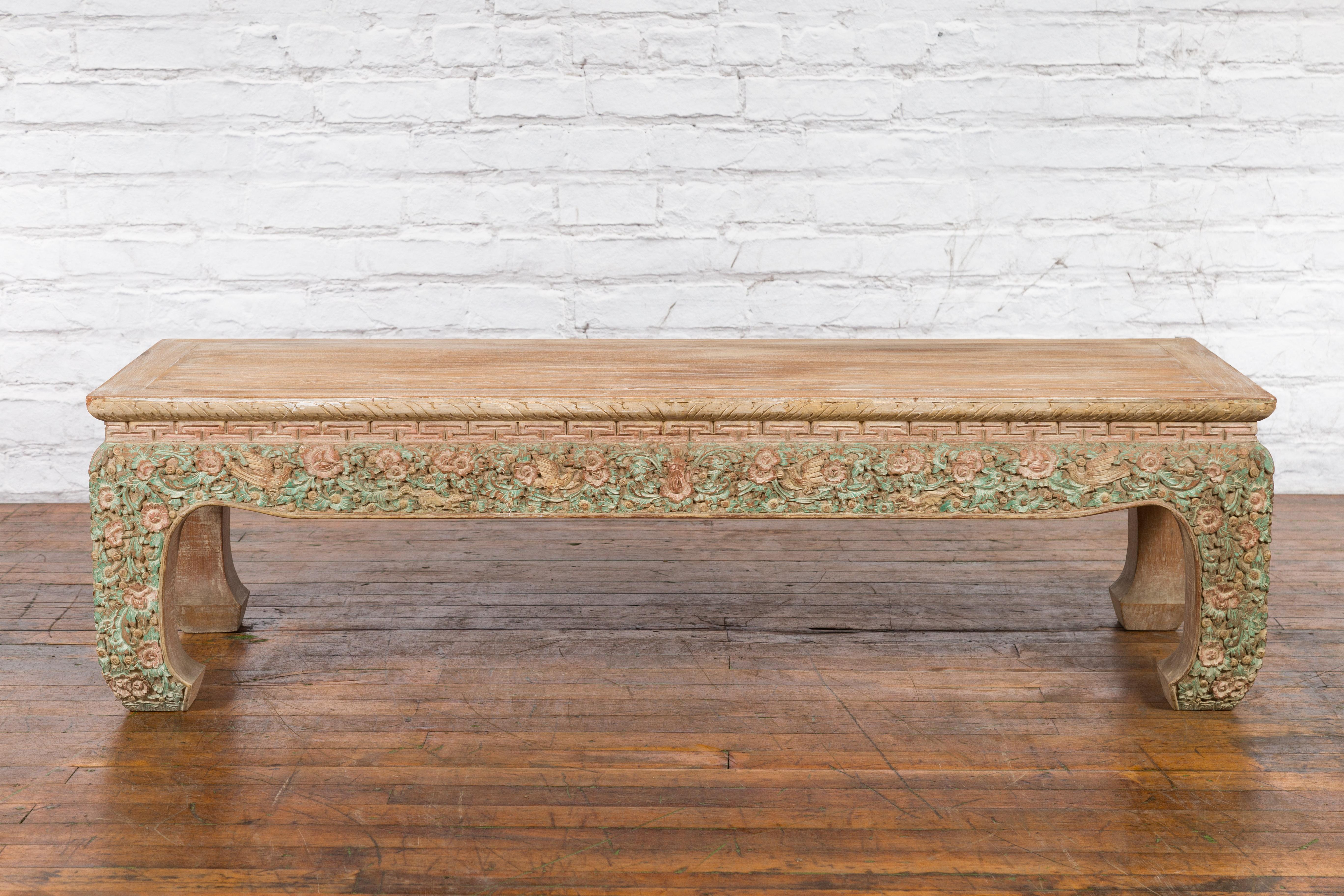 A Thai contemporary teak wood coffee table with carved floral motifs and painted accents. Created in Thailand, this teak wood coffee features a rectangular waisted top with central board, sitting above an apron carved with delicate meander motifs.