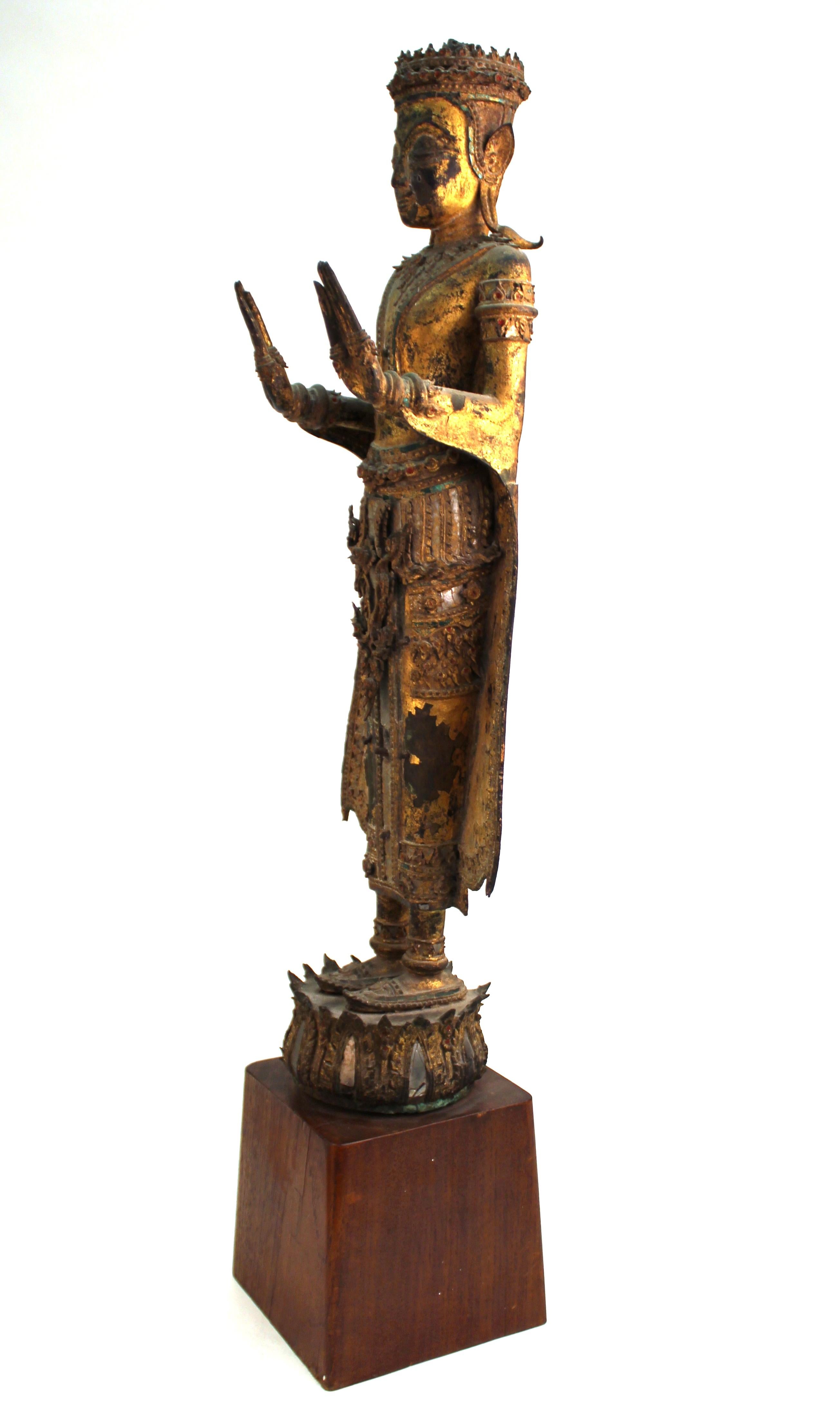 Thai antique gilded and mirrored bronze Buddha statue from the 18th century, atop a wooden base. The Buddha is wearing elaborately detailed ceremonial garments and adornments and is standing atop a lotus base. In great antique condition with