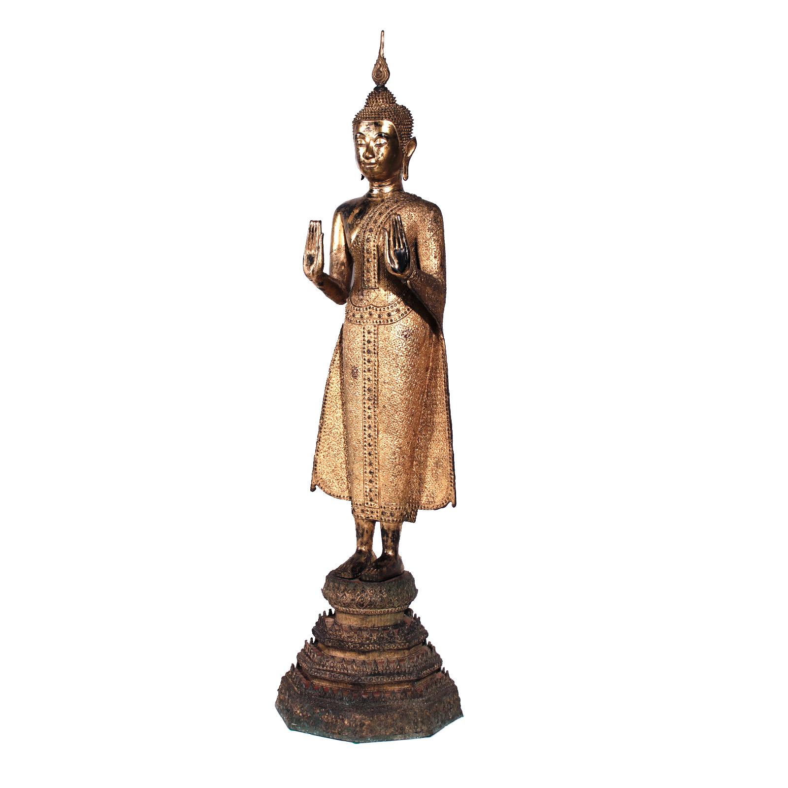 Thai gilt bronze figure of a standing Buddha, depicted upright with both hands extended forward and slightly up from the elbow, wrists bent backward, palms face forward with the long elegant fingers of the same length, reaching towards the heavens,