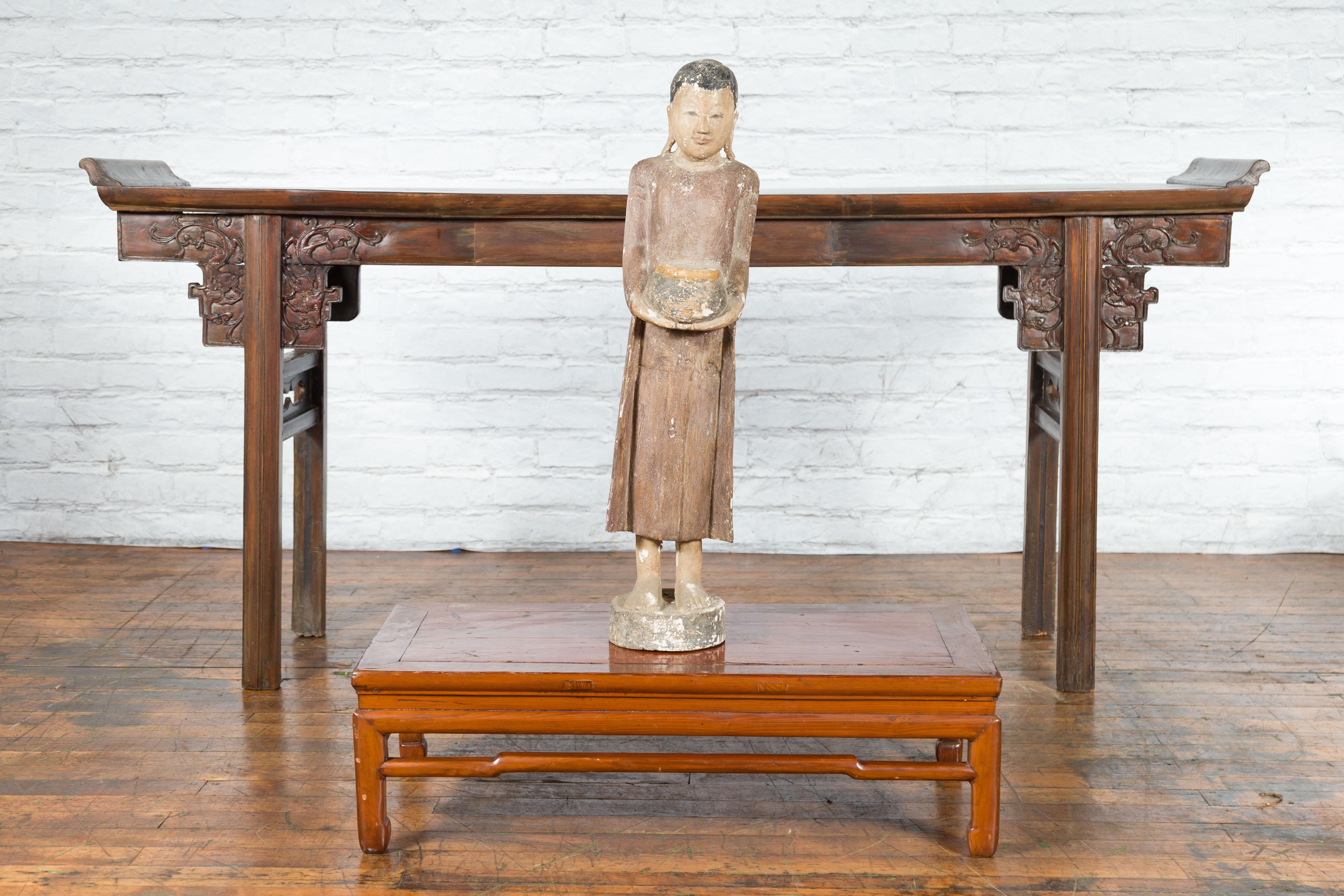 A Thai hand-carved wooden standing monk sculpture from the late 20th century, holding an offering bowl. Created in Thailand during the second half of the 20th century, this wooden sculpture depicts a Buddhist monk wearing a simple robe and standing