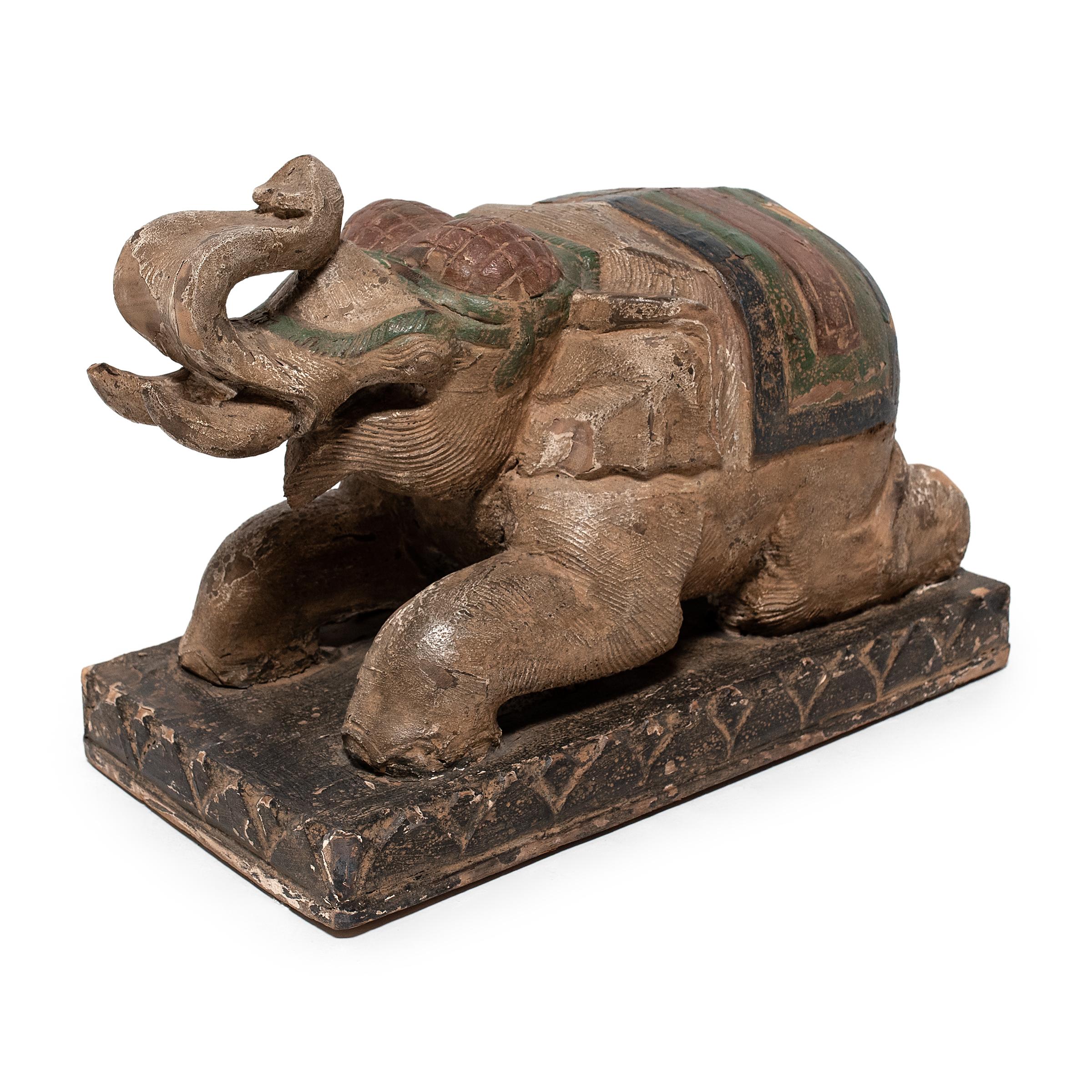 This reclining elephant was crafted in Thailand in the mid-20th century and has a carved wooden substrate with a polychrome finish. The elephant is depicted lying on its knees with its trunk in the air, a posture to allow a rider to mount its back.