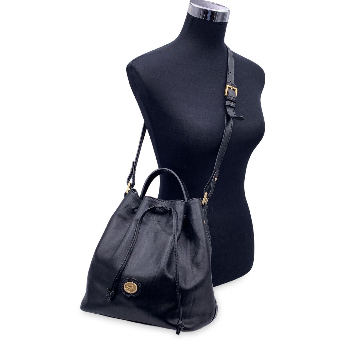 Vintage the Bridge bucket bag crafted in black leather. Drawstring closure on top. Black fabric lining. 2 main compartments with 1 middle zip section. 1 side zip pocket inside. Gold metal The Bridge logo tab on the front. Top carry handle and