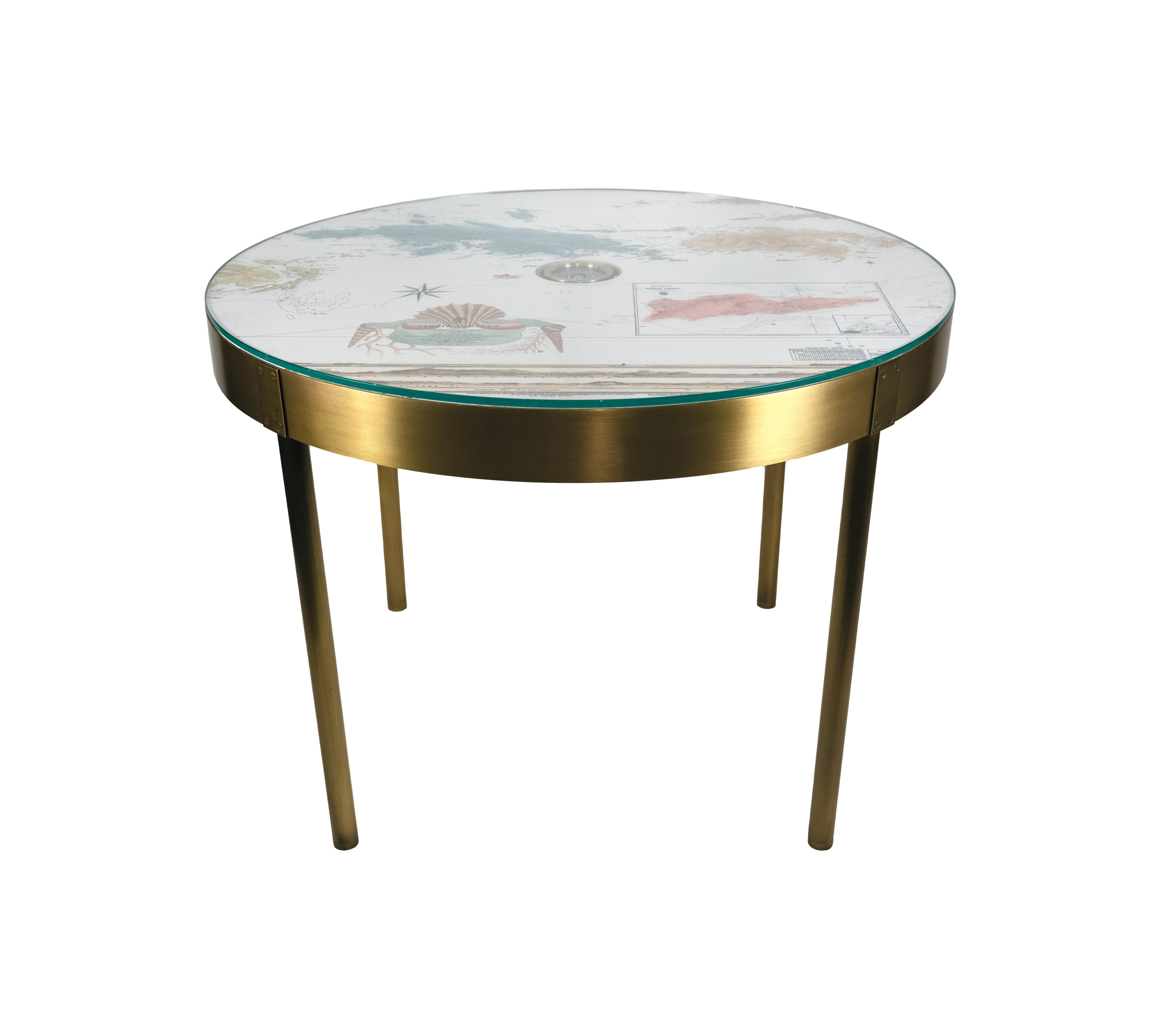 A round card table with a brass frame and printed top featuring a custom map of the US Virgin Islands, inspired by archival images. Featuring a brass compass suspended in the center, and covered with a round piece of low-iron glass. Available with
