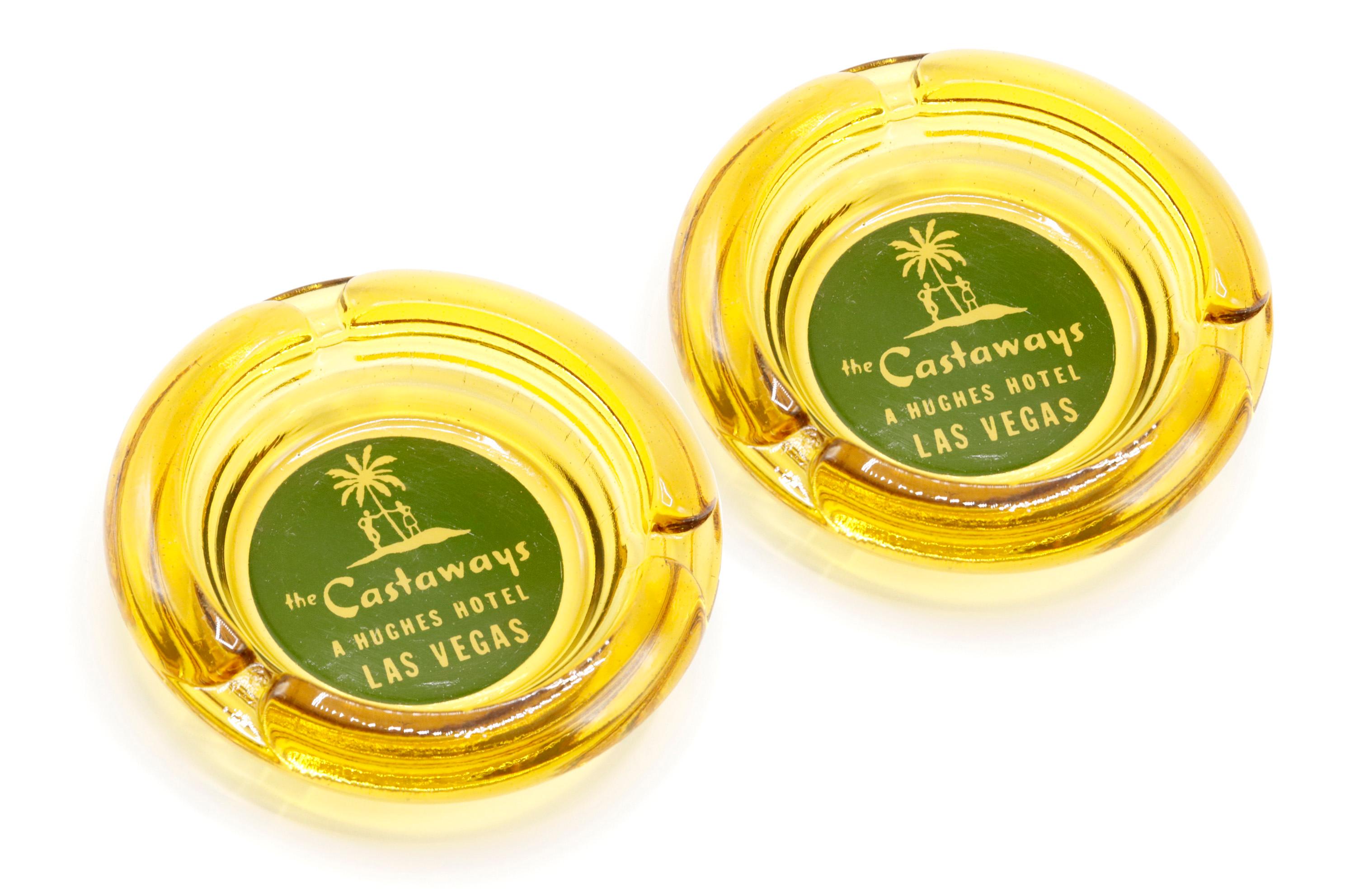 A pair of yellow glass ashtrays from The Castaways Hotel, a hotel and casino in Paradise, Las Vegas, Nevada. The center is printed with a green circle branded with the Castaways logo. The hotel, permanently closed in 1987, was opened in 1963 and