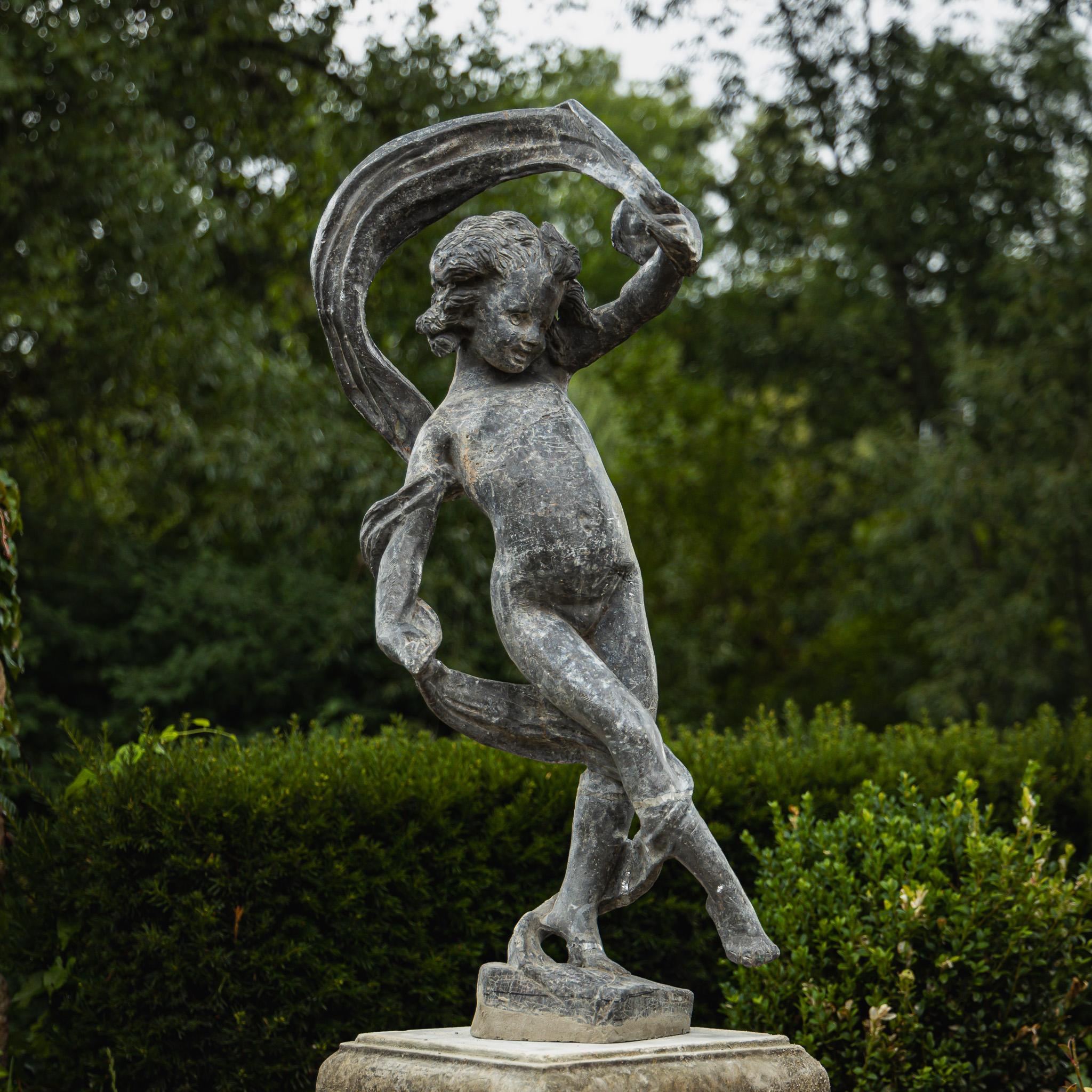 Gartenfigur eines tanzenden Mädchens aus Blei. Das Mädchen ist mit einem Schal abgebildet, der sich um ihren Körper windet und vom Wind aufgewirbelt wird. Die Bleiskulptur ist ein Klassiker und bekannt aus englischen Parks. Es ist teilweise