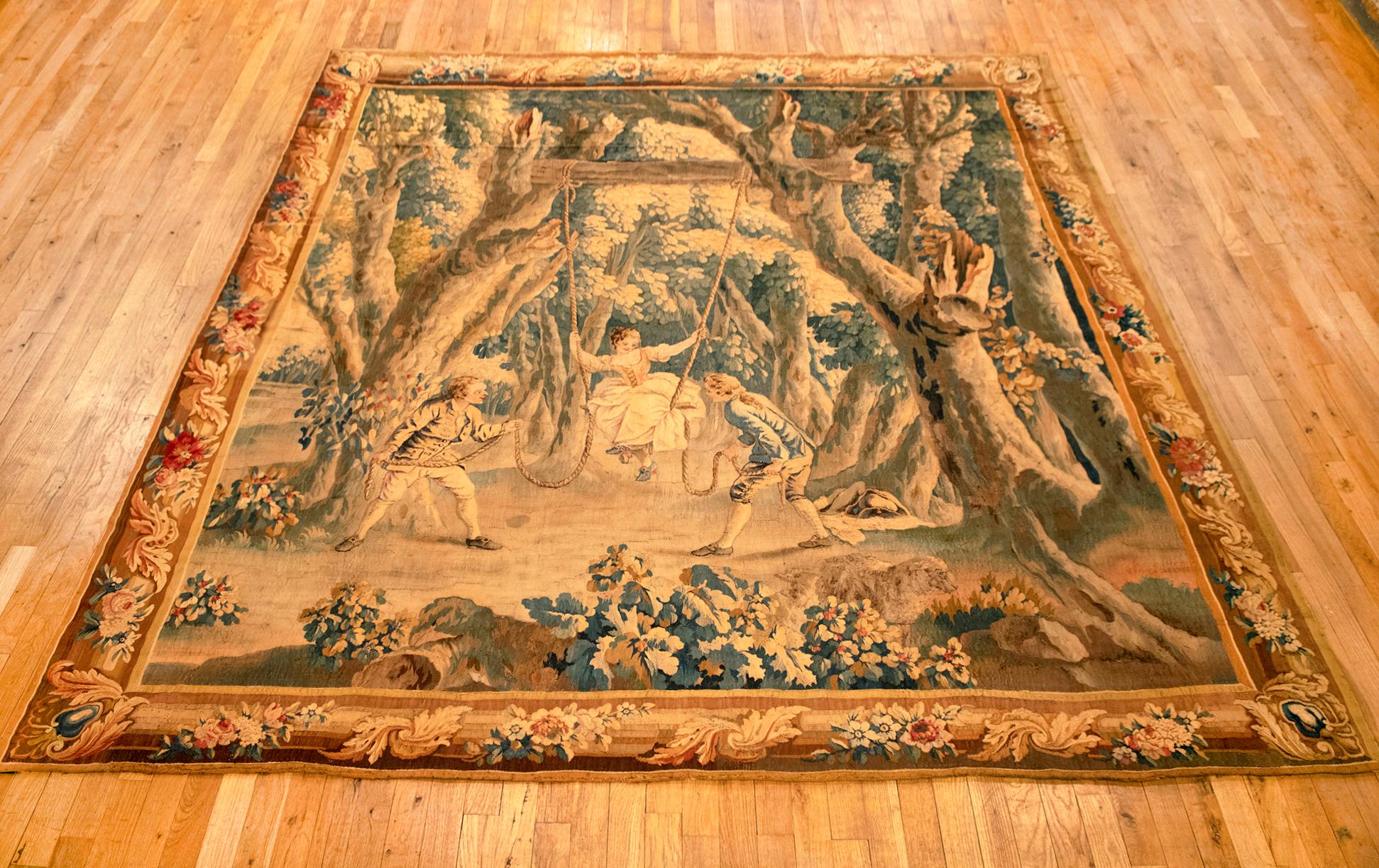 A French tapestry from the end of the 17th century, incorporating verdure and rustic elements, with a romantic scene on the foreground of two young men pulling a young girl's swing. The scene takes place in a woodland environment, with trees in the