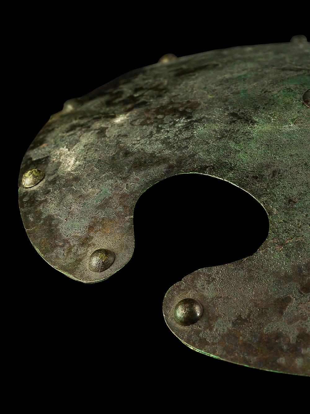 An unusual and remarkably well-preserved bronze shield, most likely of Persian origin. Oval and slightly convex, consisting of bronze sheets riveted together, with the rivet heads visible as a decorative effect round the rim and a central studded