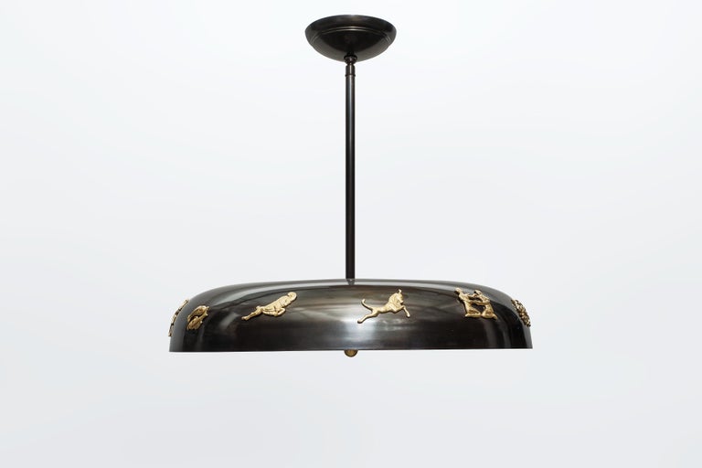 A pendant light fixture featuring gilt bronze astrology zodiac motifs mounted on a torus shaped, patinated frame. This unique and elegant fixture, inspired by the Swedish Grace period, is suspended from a single rod, with a frosted glass shade