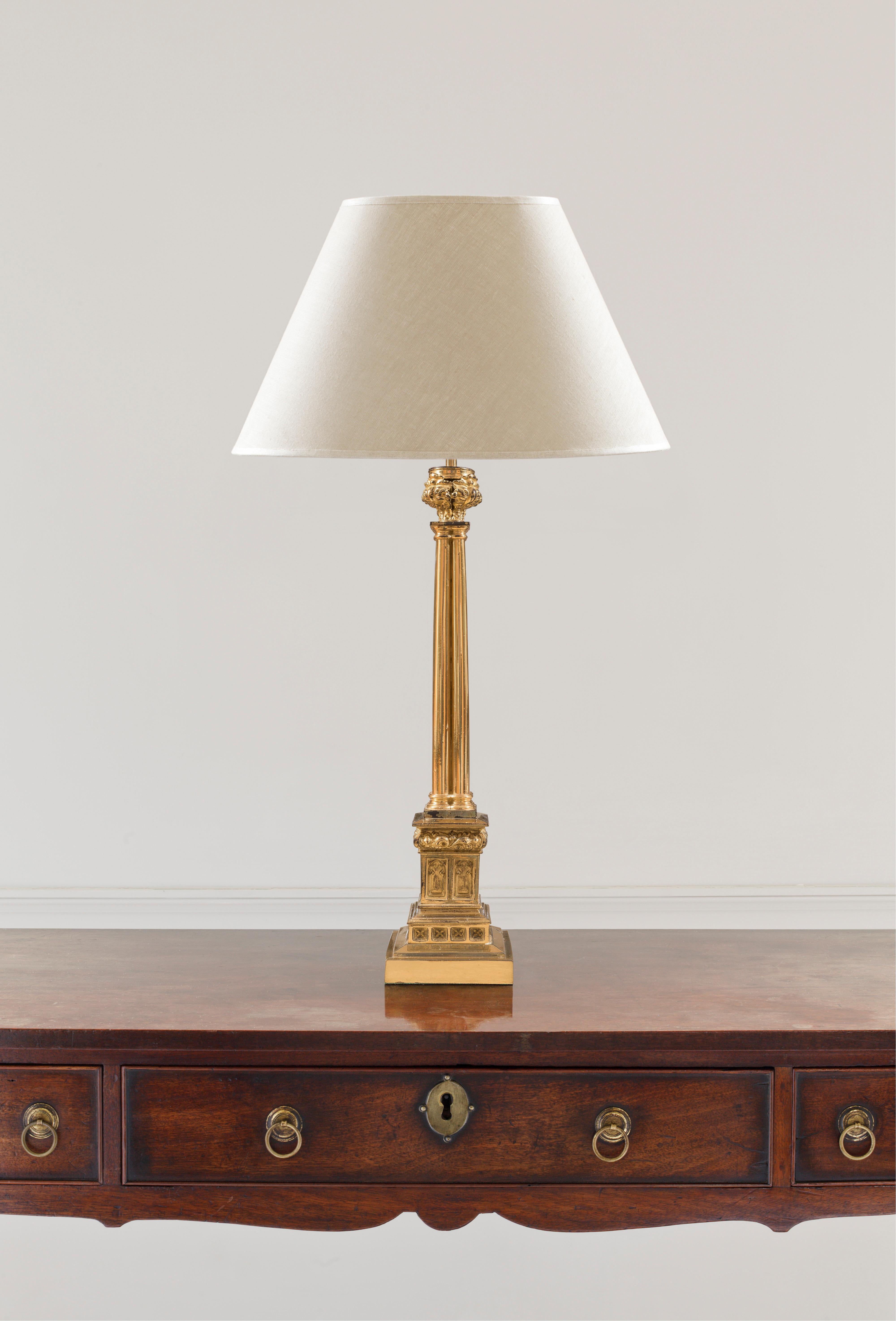 This polished bronze lamp of architectural form incorporates a foliate cap above a cathedral-like cluster of four tapering columns terminating in a detailed cast bronze pedestal, decorated in the Regency Gothic taste with stylized leaves, blind