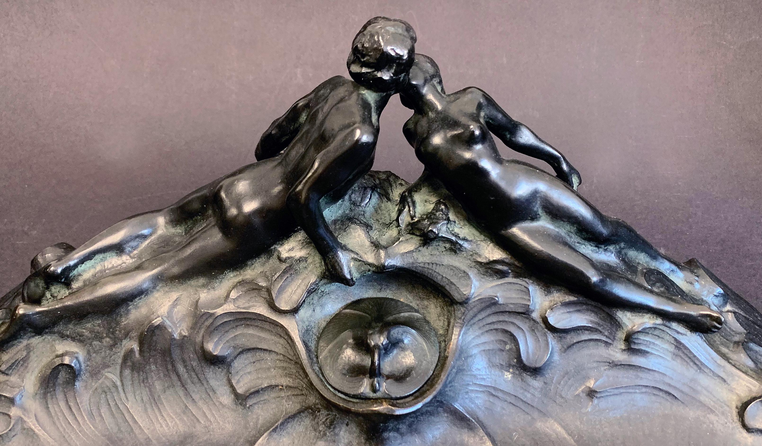 Rare and striking, this large, bronze desk accessory, a combination inkwell and large pen tray, depicts nude male and female figures, shown at the crest of a wave, in the midst of a gentle kiss. This sculpture puts the couple in an aquatic setting,