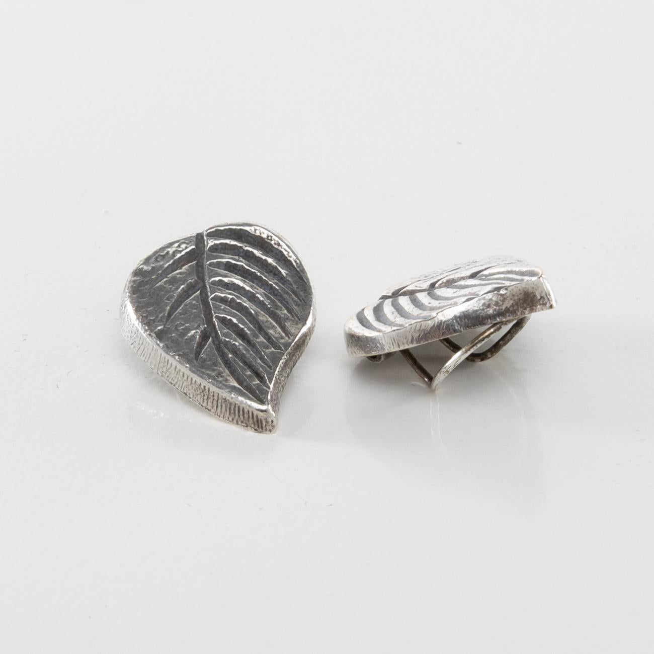 This pair of silvered bronze earrings represents two tree leaves. Each earring is different. It has a clip fastening system. 
 
The left earring is signed by stamping “L.V.”. Manufactured in the 1960s, this piece of jewellery is in very good