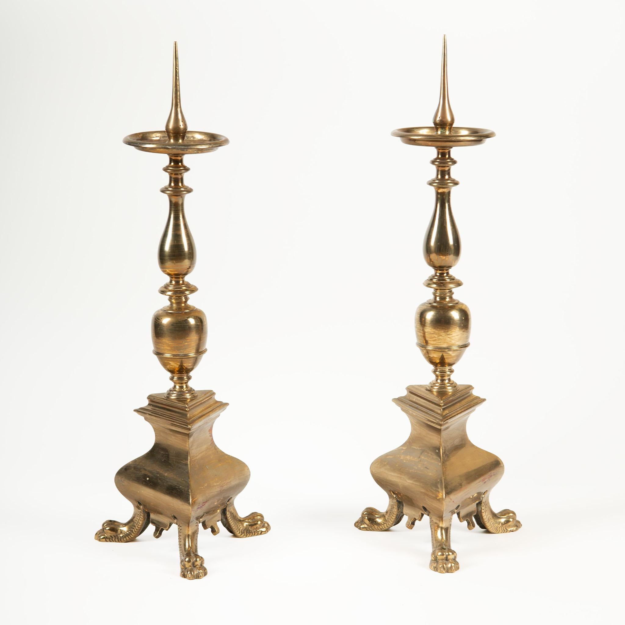Pair of Brass Altar Pillar Candle Holders, early 20th century, featuring an elegant turned baluster stem above a wide drip pan with central pricket, designed for large pillar candles. The bases are boldly formed with curvilinear sides, raised on