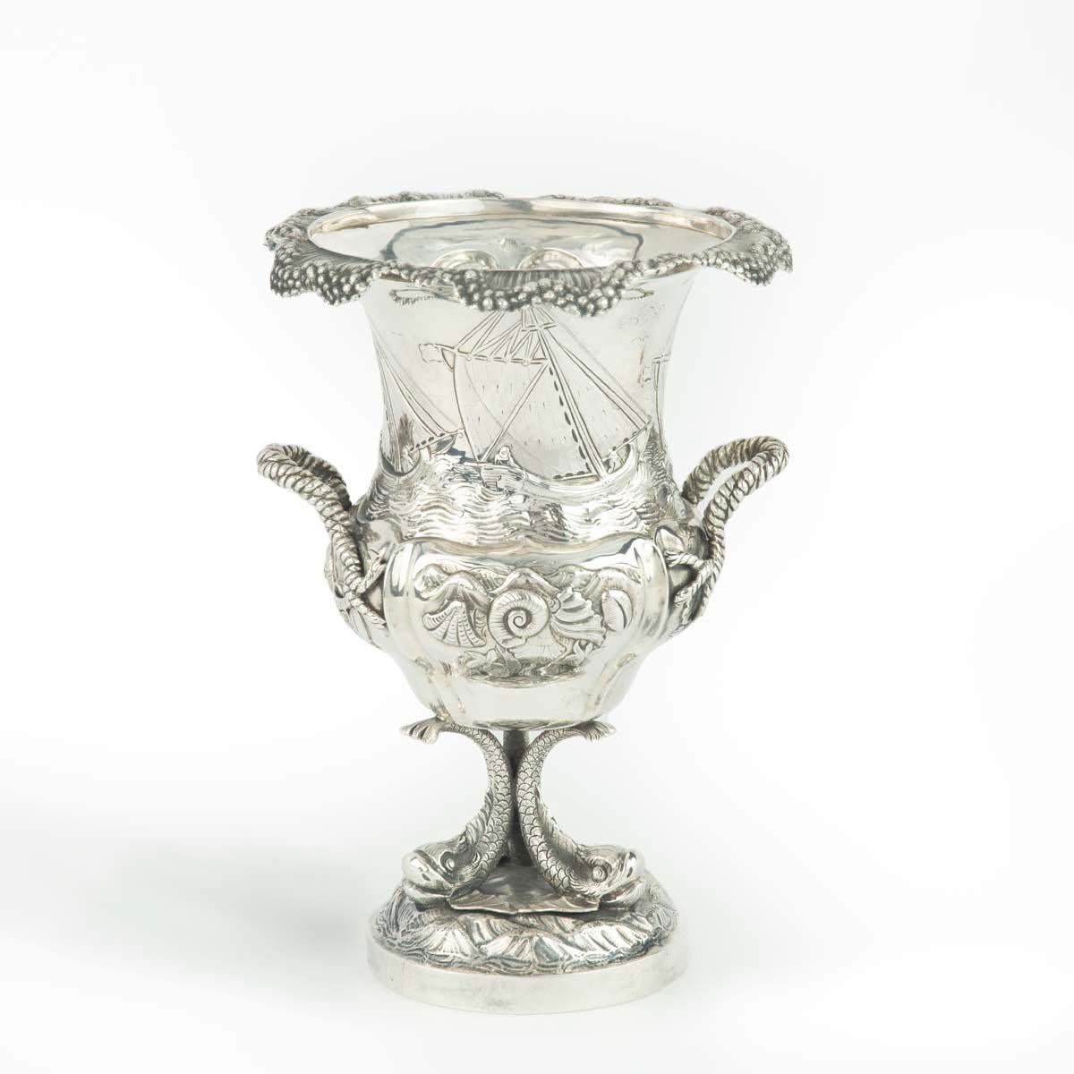 This ornate repoussé silver trophy is in the form of a handled cup with an everted rim and tripod dolphin base.  The high relief decoration comprises three gaff rigged yachts racing, above a frieze of shells against waves.  There is a C-scroll