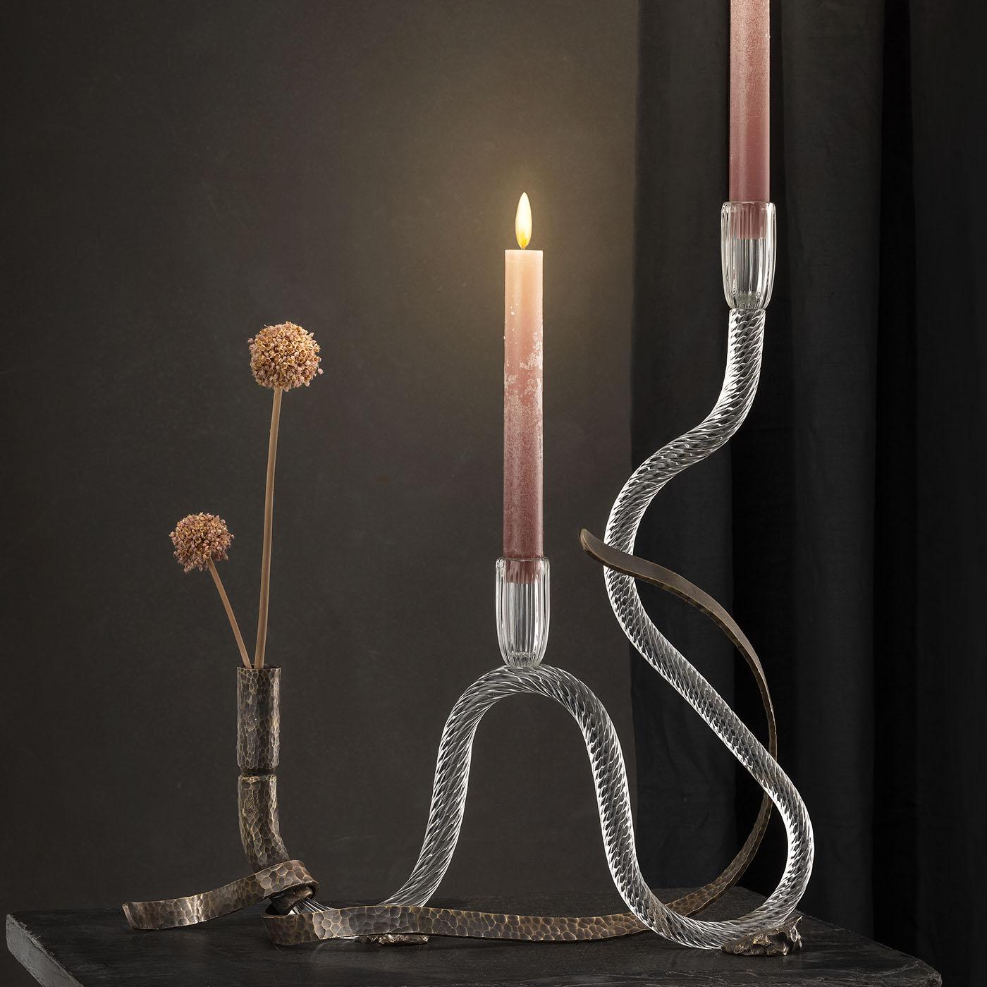 The One curved Murano glass candle holder features a sinuous, sculptural form that blends hand-hammered brass with mouth-blown glass. The design offers a dynamic interplay between textured metal and smooth transparency, emphasizing a refined yet