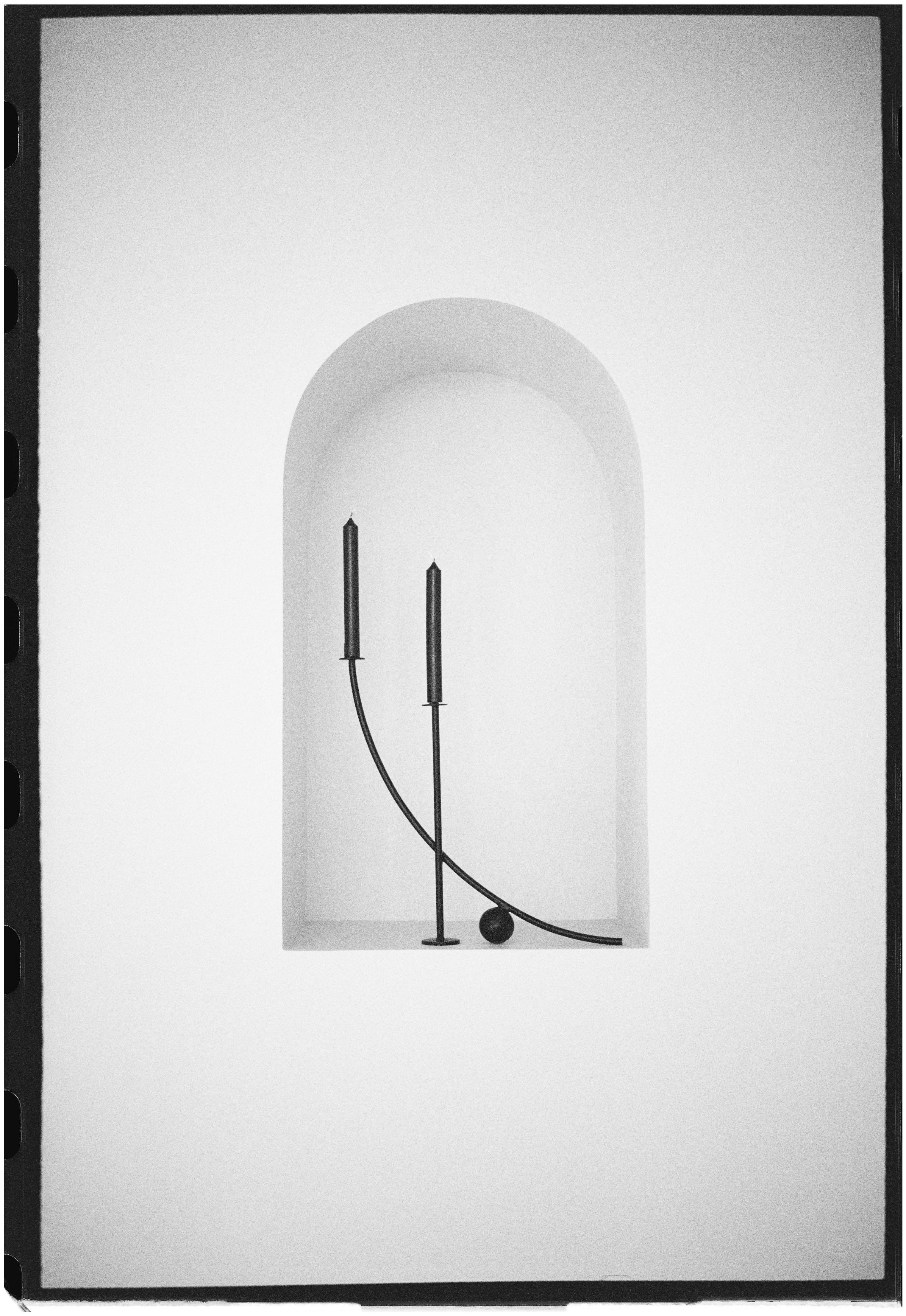 A study in balance and motion, The Orbit reimagines the traditional candleholder as sculptural form. Two slender arms arc upward from a single axis, each bearing a minimalist taper. One curve anchors itself in precision, while the other completes a