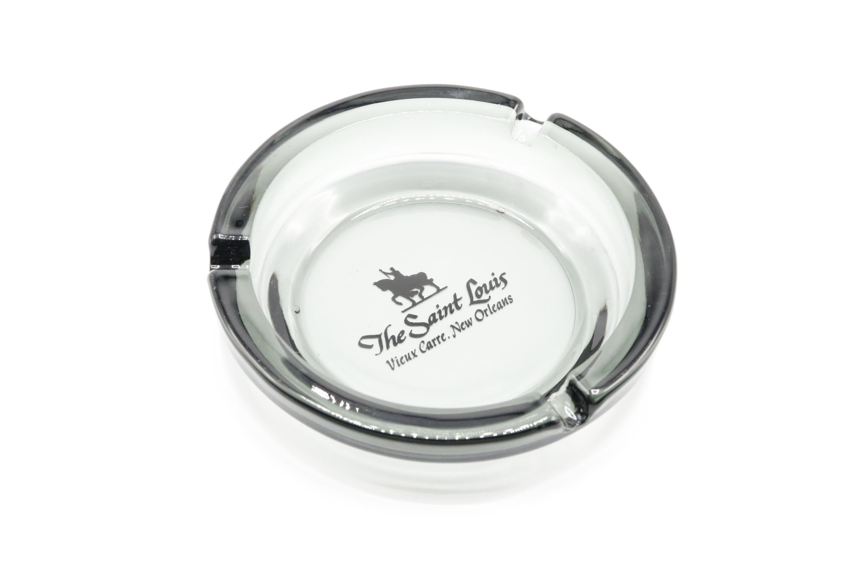 A gray tinted glass ashtray from The Saint Louis Hotel, now the Hotel Mazarin, in the French Quarter, New Orleans. The center is printed with their logo in black and reads “The Saint Louis, Vieux Carre, New Orleans”. 