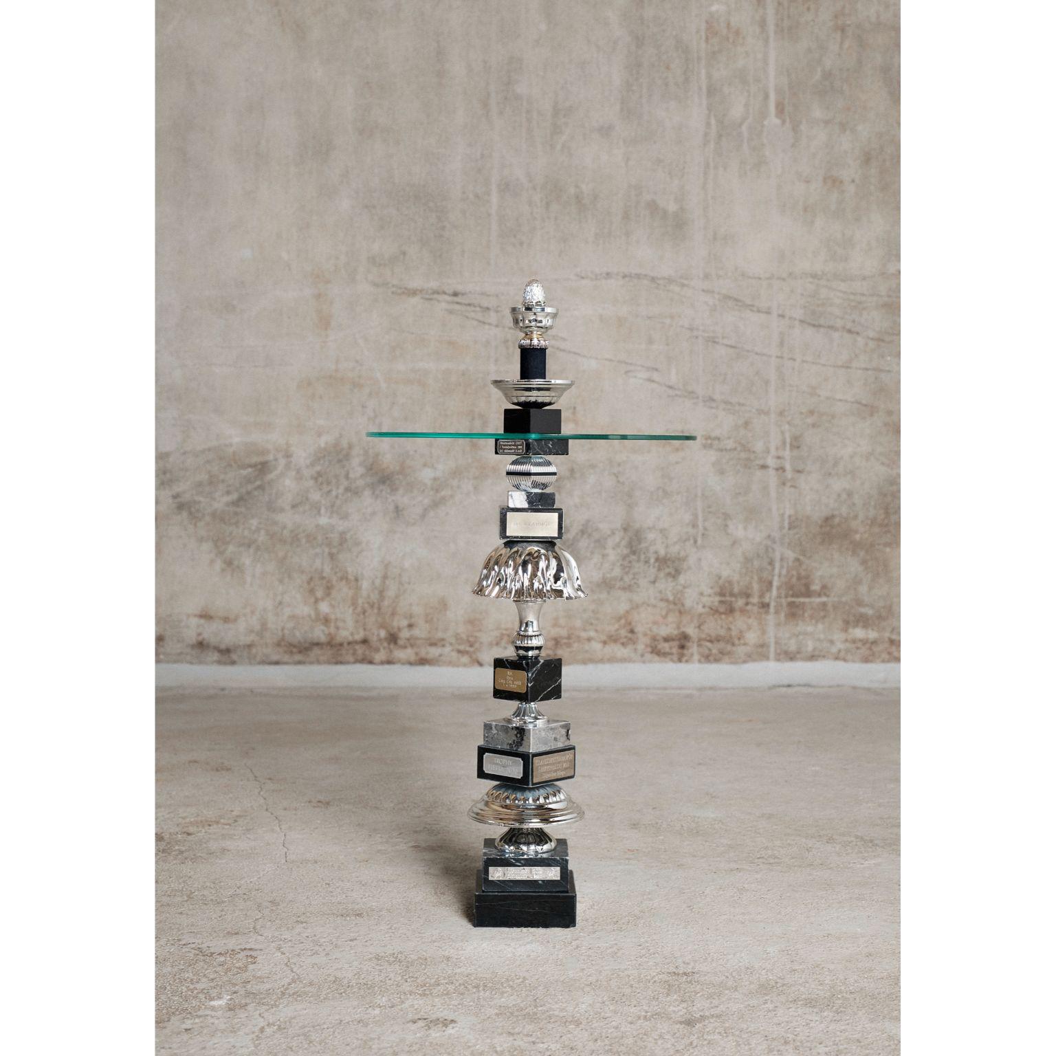 The silver pine cone table by Flétta
Dimensions: D40 x H76 cm
Materials: Black/silver/grey marble, glass.

Trophy is a collection of tables, lights, flowerpots and shelves made of old trophies collected from athletes and sports clubs in Iceland.