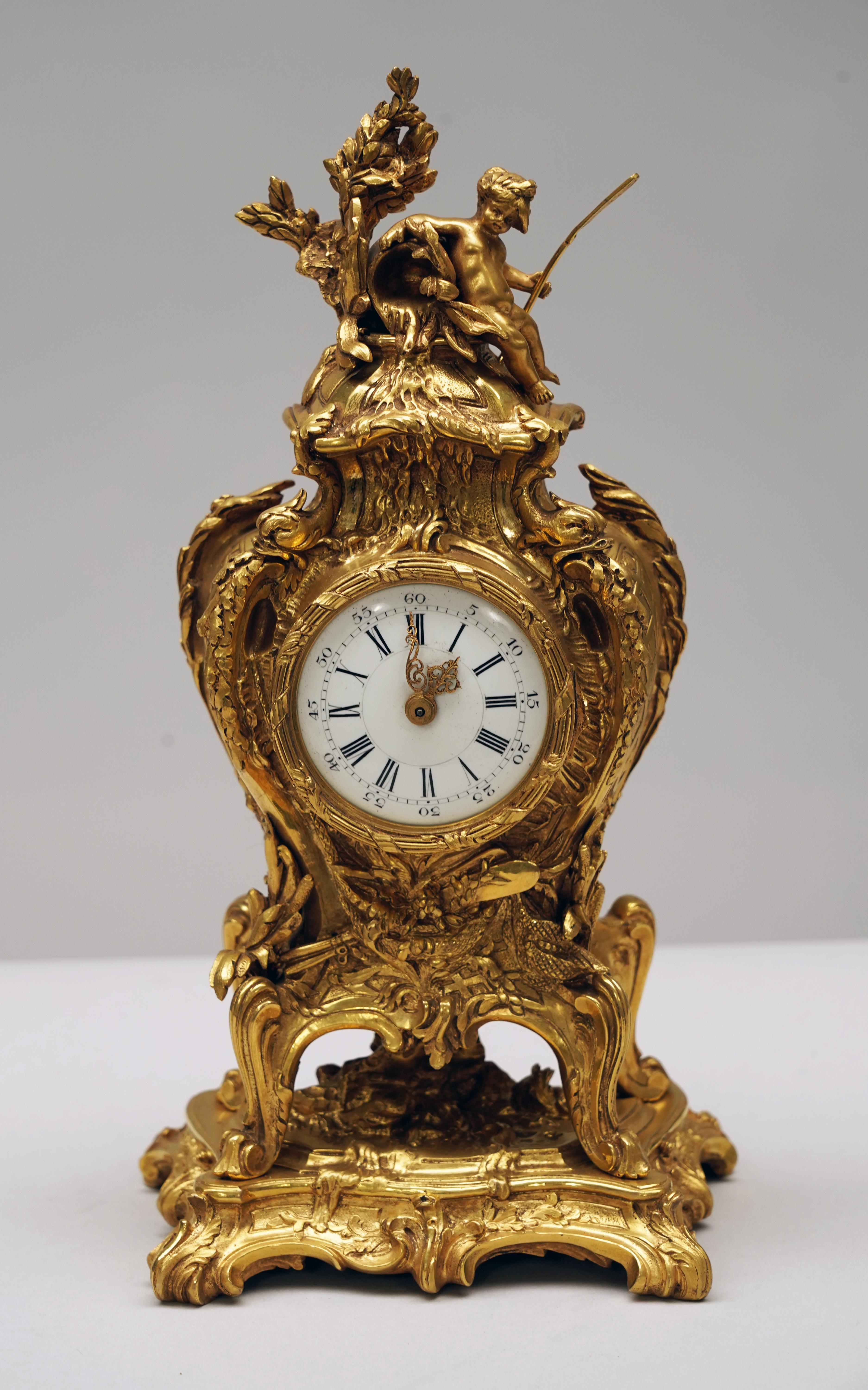 Charming clock in the shape of a Rocaille-inspired cartel in chiseled and gilded bronze. Scalloped marine-inspired decor representing a trophy with oars, dolphins, reeds and flowers on a trellis background. The white enamel dial is surmounted by an
