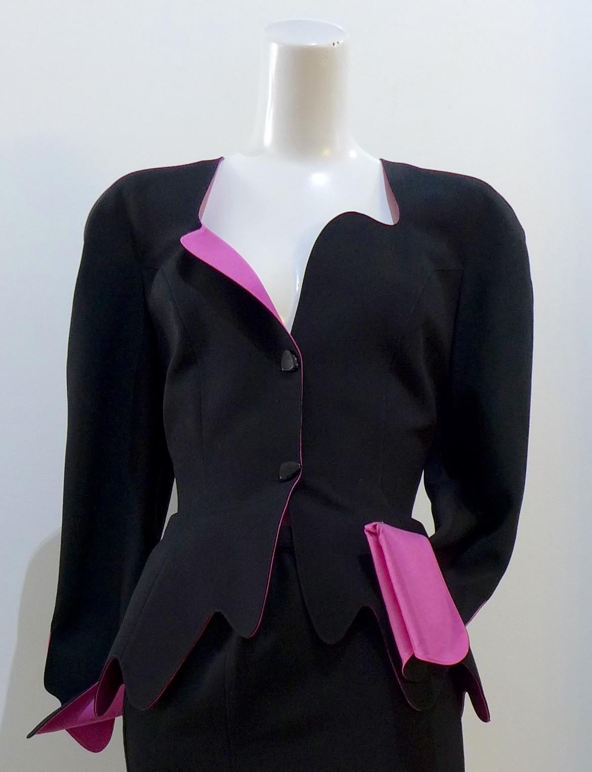 This THIERRY MUGLER skirt suit is composed of a black wool fabric with bright pink lining and cuff. Features a classic Mugler silhouette with nipped waist and curved bust-line to neckline design. The jacket has front snap closures, rounded