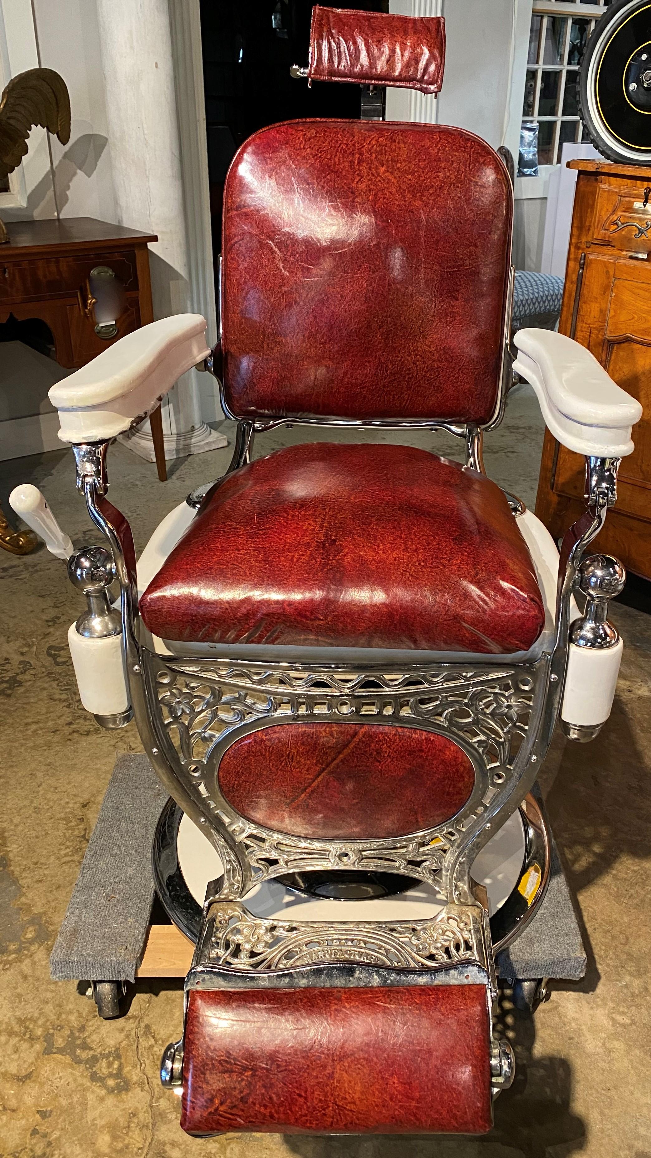 A fine example of a heavy chromed, porcelained, and scrolled metal frame barber chair by Theo A. Kochs, Chicago, with porcelain arms, red leather seat, back, headrest, and footrest, adjustable back and swivel seat height (hydraulic pump handle on