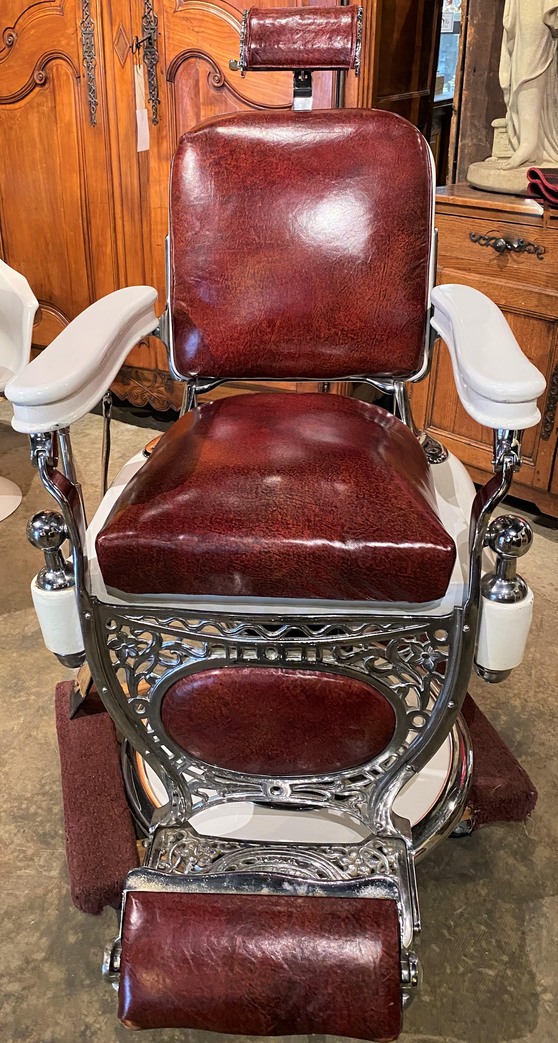 A fine example of a heavy chromed, porcelained, and scrolled metal frame barber chair by Theo A. Kochs, Chicago, with porcelain arms, red seat, back, headrest, and footrest, adjustable back and swivel seat height (hydraulic pump handle on side), and