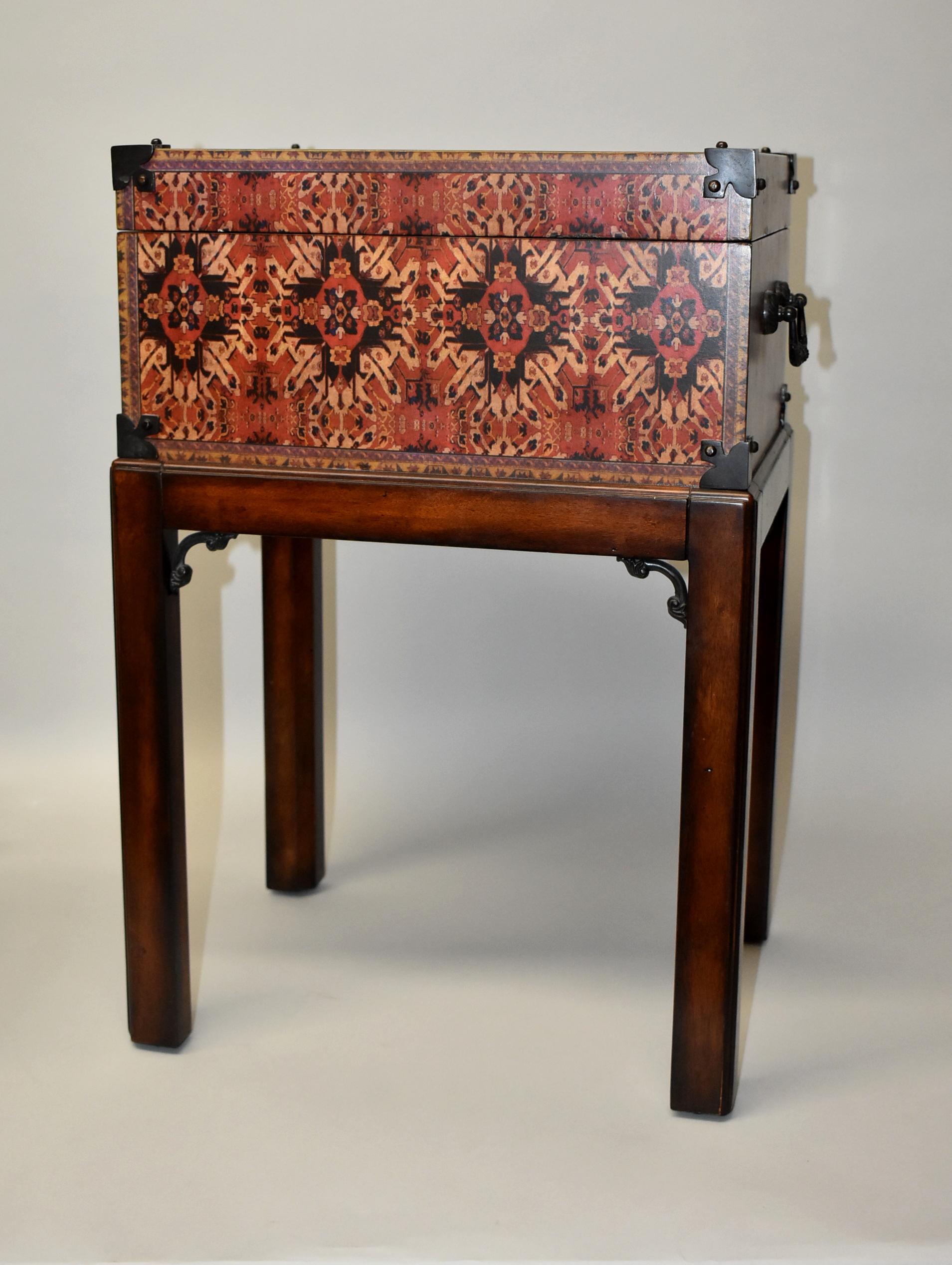 Theodore Alexander wood box decorated with an oriental rug motif, metal corners, a hinged lid, and ornate brass handles. The box sits on a stand with decorative leg supports. 24 1/2