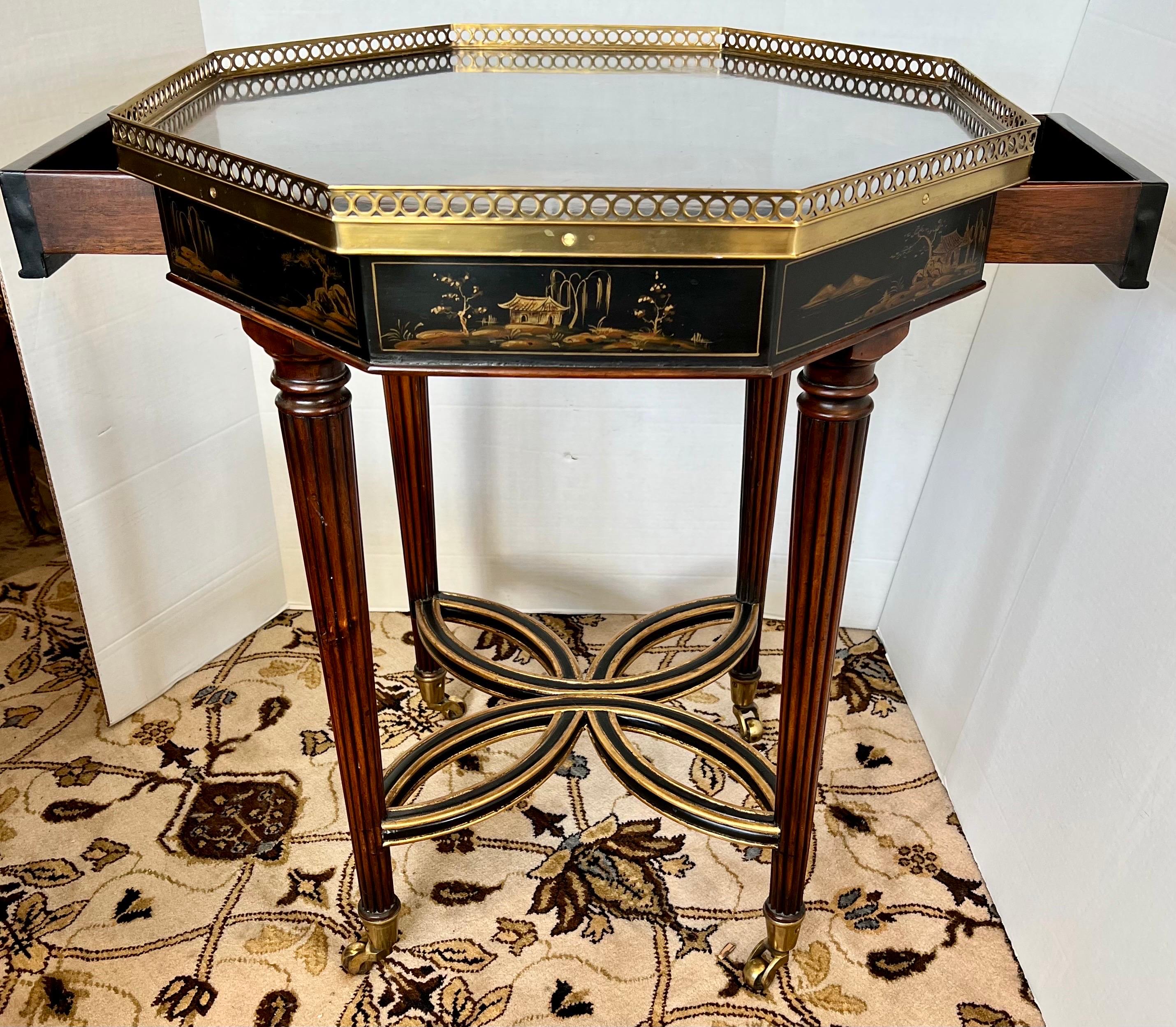 Tunning chinoiserie black lacquered octagonal table with painted gold scenery on the top and sides. Two drawers on either side and pierced brass gallery. Carved fluted legs with brass castors for ease of movement. Theodore Alexander plaque inside