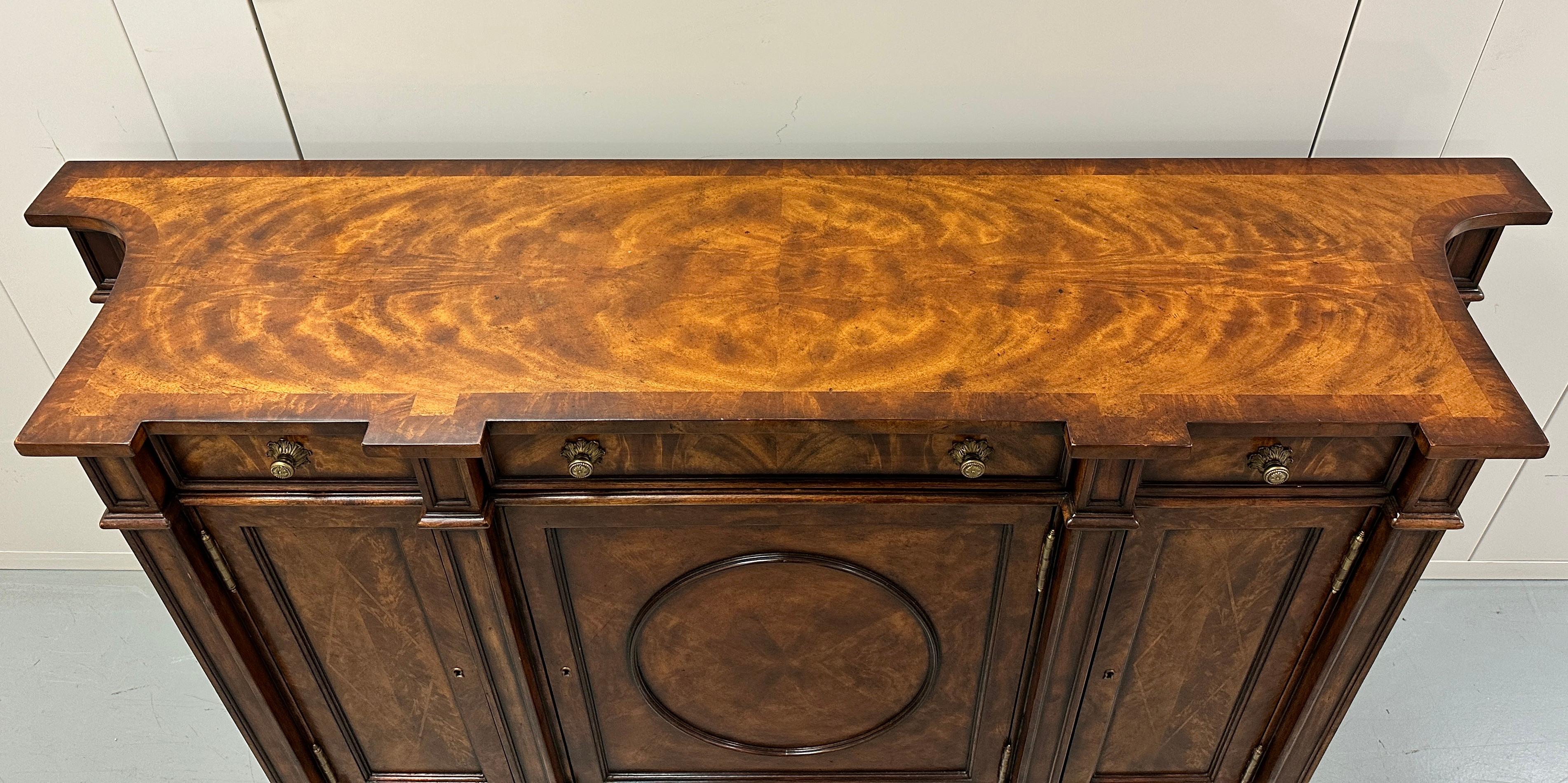A fine mahogany, flame veneered and burl banded side cabinet, with three frieze drawers above corresponding oval panel and lozenge inlaid doors enclosing adjustable shelves, with concave sides, on turned legs with brass cappings. Replica collection.