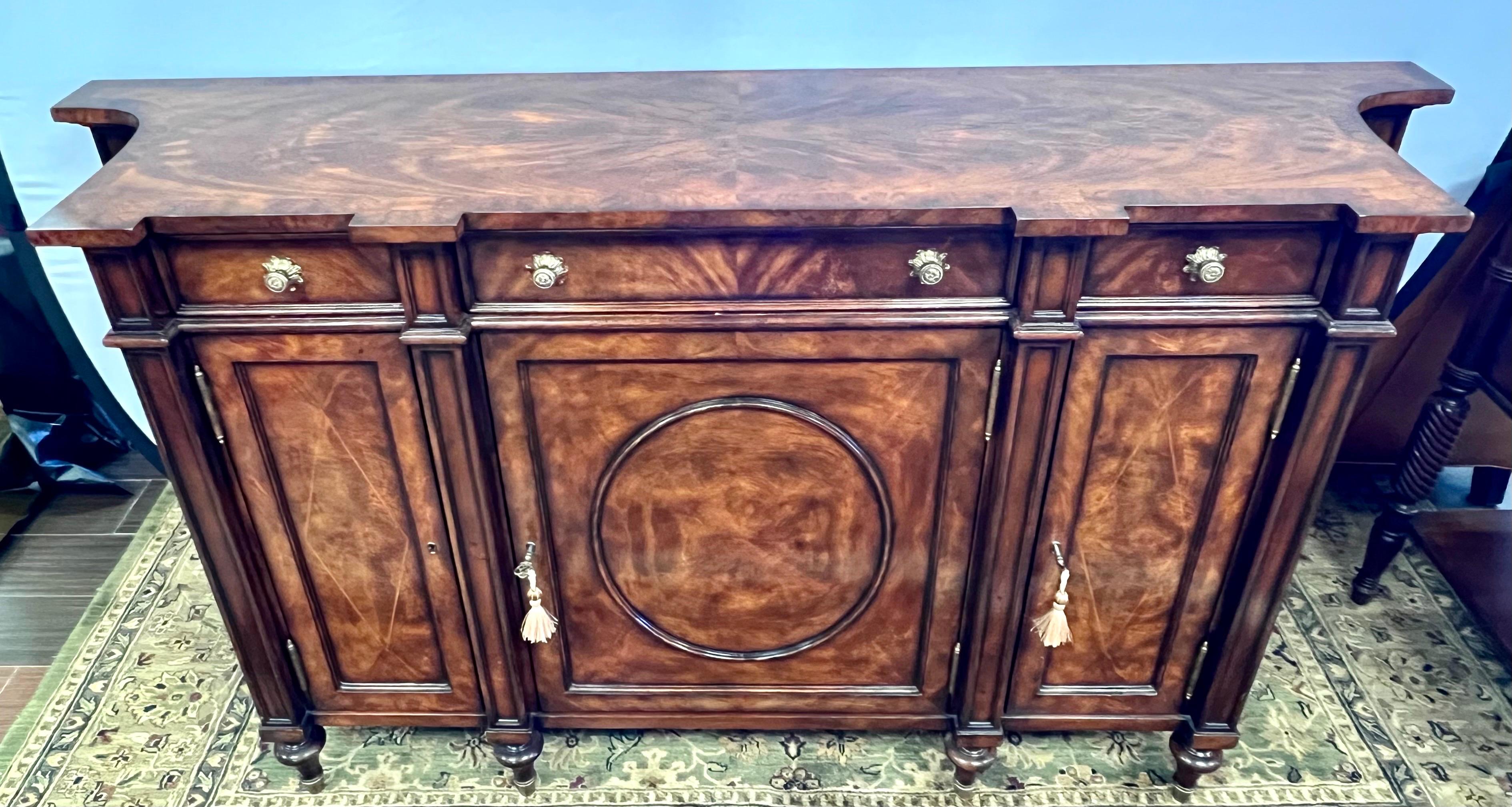 Beautiful Theodore Alexander mahogany, flame veneered and burl banded sideboard, with three drawers above corresponding oval panel and inlaid doors that open to adjustable shelves, with concave sides, on turned legs with brass cappings.