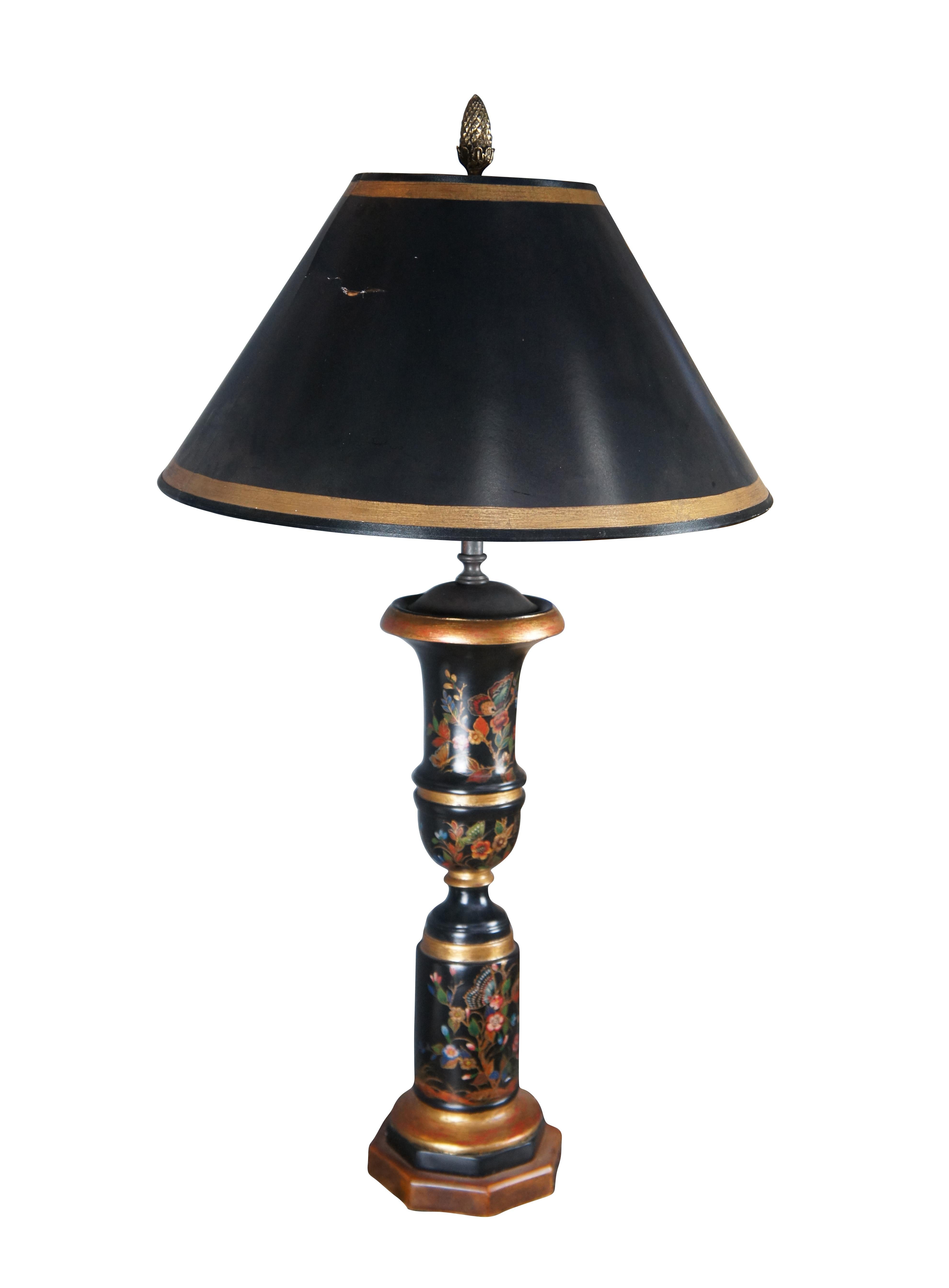 A beautiful Theodore Alexander Tole style table lamp. It features a trumpet or trophy urn form with a black hand painted wooden column decorated in colorful flowers and butterflies. The lamp is accented in gold and has a black tapered shade with