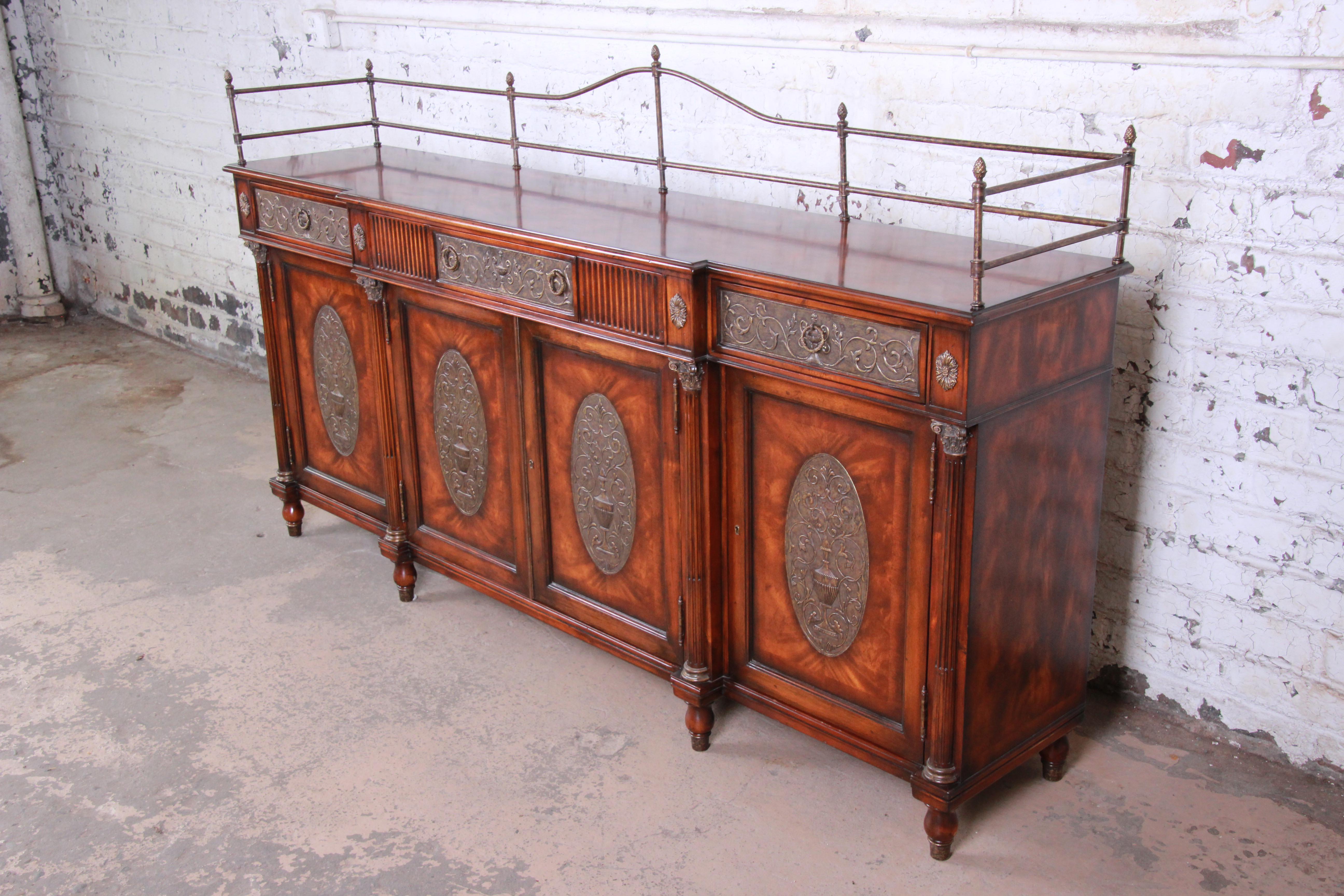 An exceptional and monumental Regency style sideboard buffet or bar cabinet by Theodore Alexander. The sideboard features gorgeous flame mahogany wood grain, Corinthian columns, and ornate brass details with a gallery top. It sits on turned legs