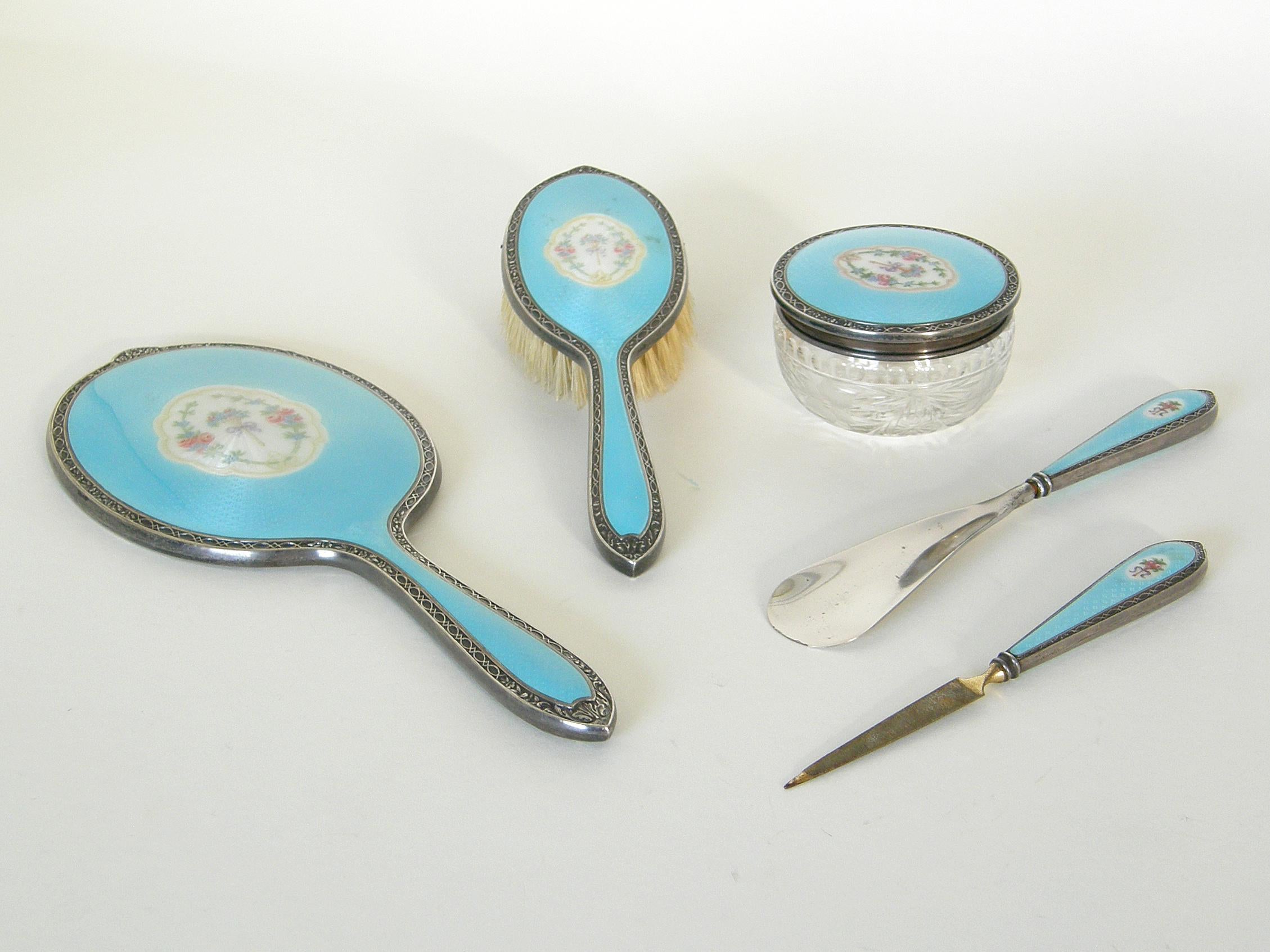 This lovely, antique vanity or dressing table set was made by Theodore W. Foster & Bro., of Providence, Rhode Island. The five-piece set is made of sterling silver with bright, baby blue guilloché enamel. The enamelwork features central medallions