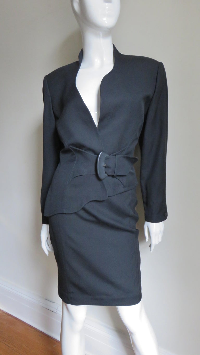 A beautiful black 2 piece skirt suit from Thierry Mugler.  The jacket is collarless with an asymmetric neckline and hem, shoulder pads and a front adjustable belt with a black buckle. The pencil skirt has a waistband with 2 black snaps, a back