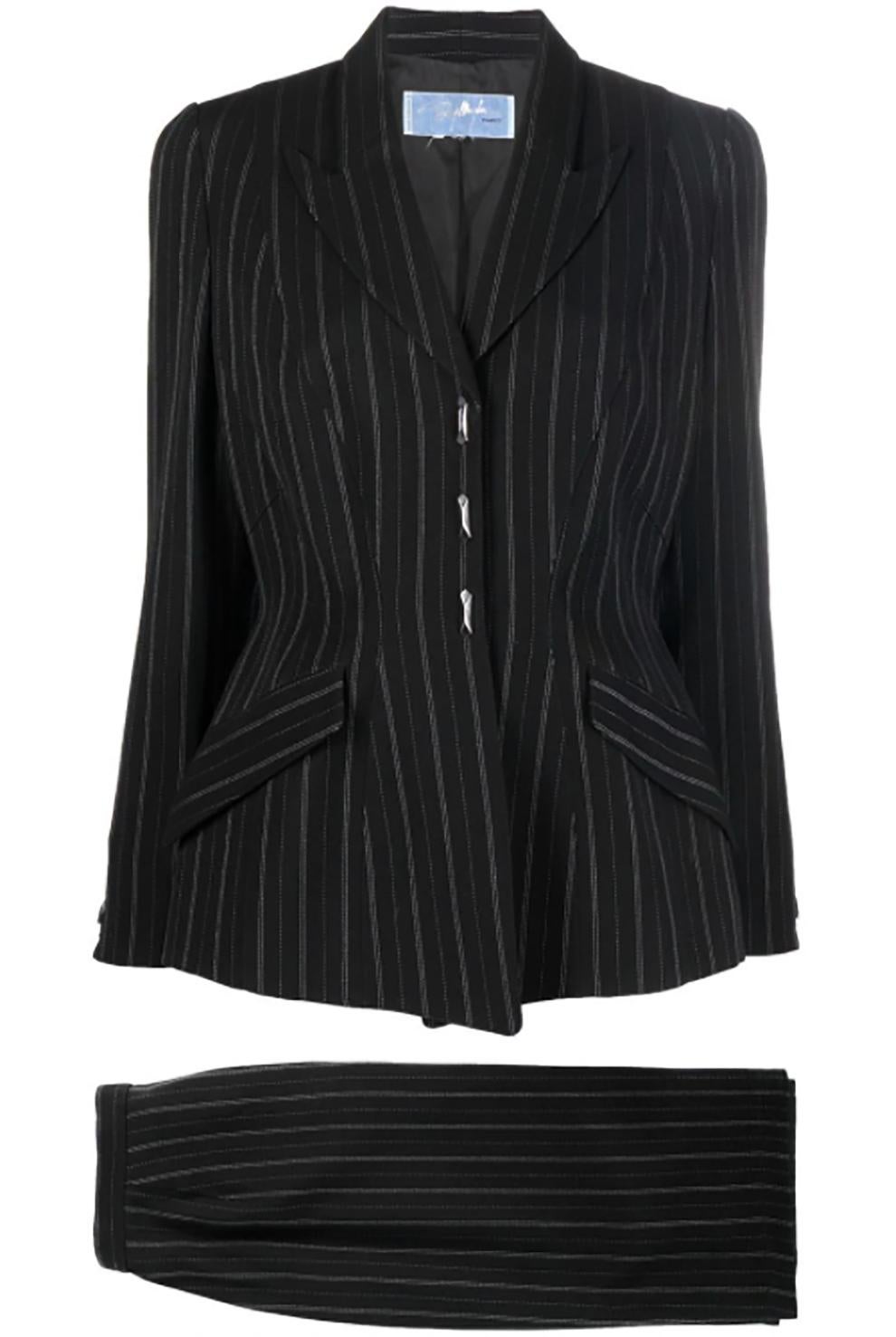 Abito nero di Thierry Mugler con motivo gessato, giacca aderente in vita con revers a lancia, polsini con bottoni, due tasche laterali con patta.
E una gonna a matita aderente con vita alta, lunghezza al ginocchio e chiusura nascosta.
Composizione: