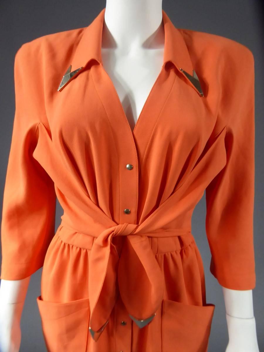 Circa 1988

France

Mid-length orange gown shoulder three quarter sleeves signed Thierry Mugler .Closes on the front with a buttonhole of ten silver snap buttons and a stitched belt on the hips ending with two silver chevrons at the ends recalling