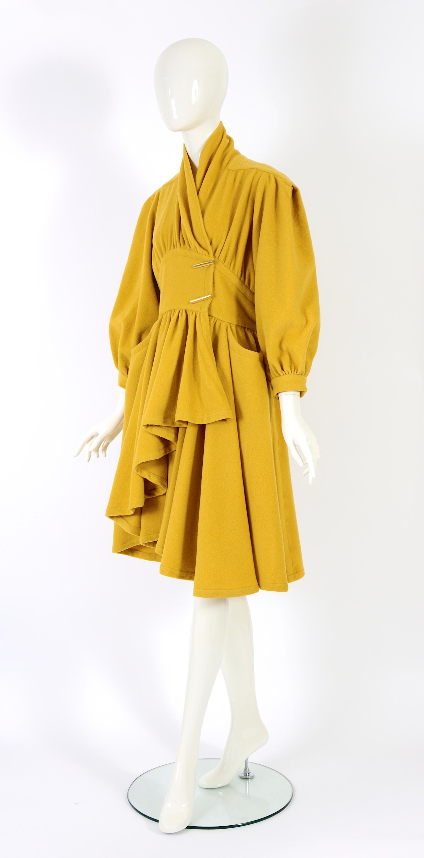 Documented Thierry Mugler Fall Winter 1983 runway, mustard yellow wool coat, with massive shoulders, graduated gathered skirt cut short at the front, broad curving waistband with two gilt bar fasteners.
Made in wool - No size label - approximately