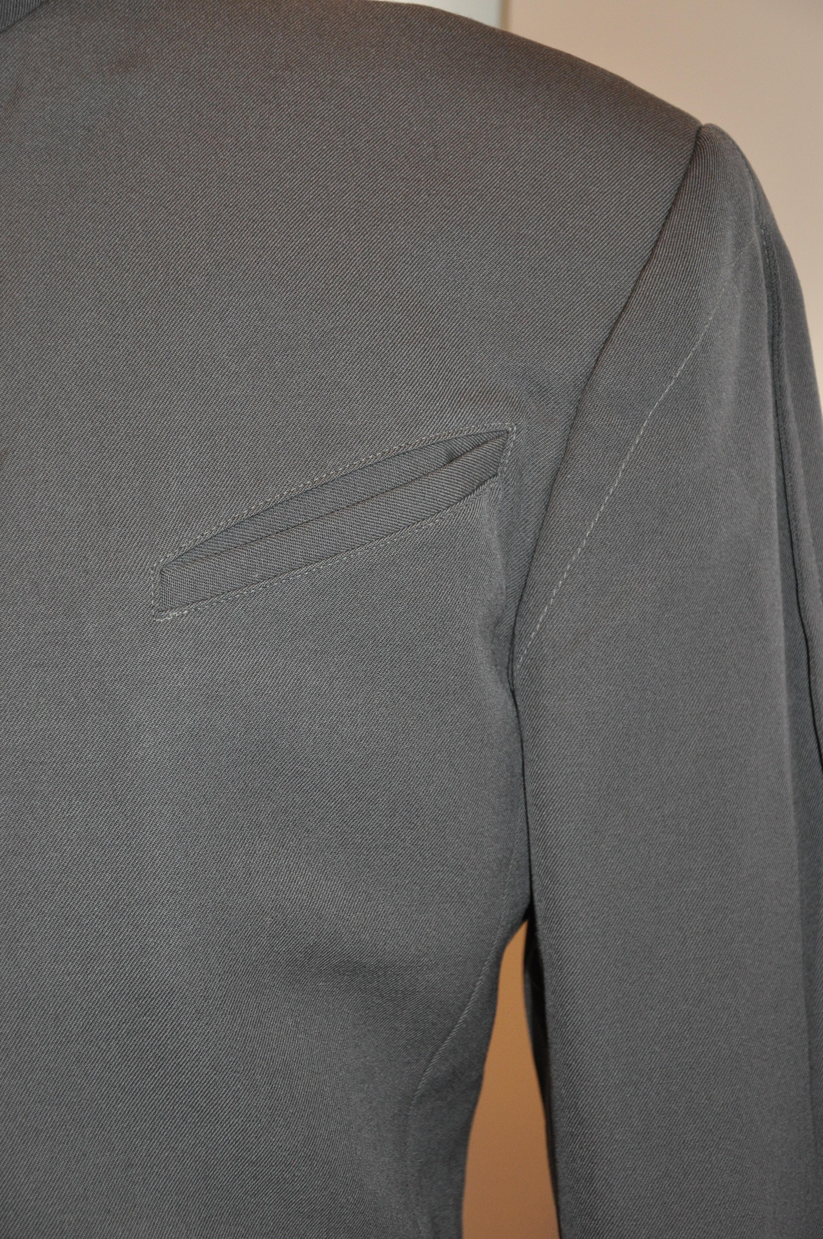        Thierry Mugler è una rara giacca iconica da uomo in lana color taupe con collo a scatto e colletto alla mandarina. La parte anteriore ha sei (6) bottoni automatici. Ci sono due tasche incastonate e due tasche incastonate all'interno. Il