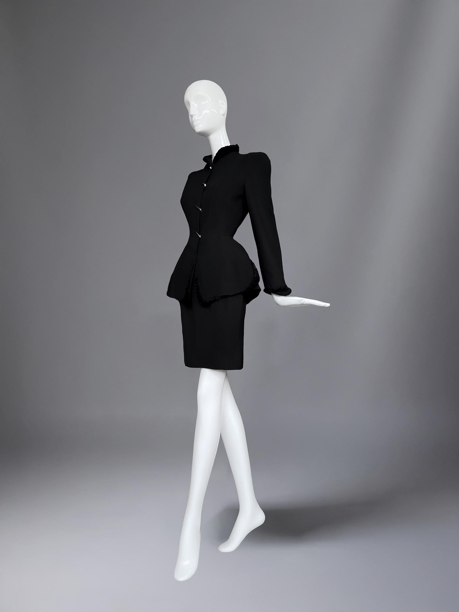 Le génie de Thierry Mugler résidait dans sa capacité à transformer le tailleur en théâtre, et ce tailleur jupe noir est un parfait exemple de sa Couture sculpturale. La silhouette est du pur Mugler : une veste ajustée qui s'évase en un péplum