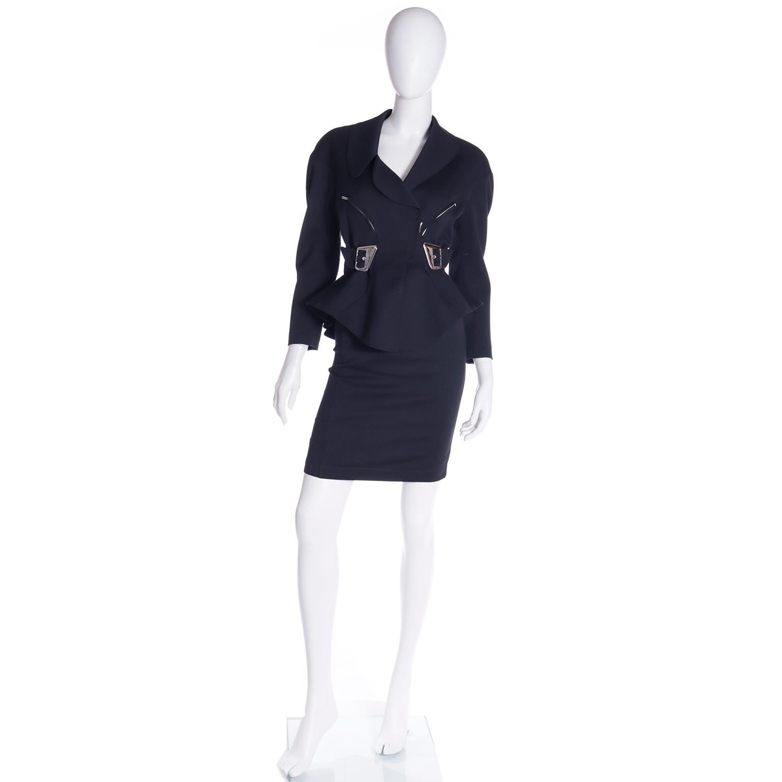 Questo tailleur a due pezzi di Thierry Mugler dei primi anni '90 è in lana nera e presenta grandi fibbie d'argento. La giacca ha un colletto asimmetrico, una cerniera sul davanti per la chiusura e due tasche con zip. La giacca è fortemente