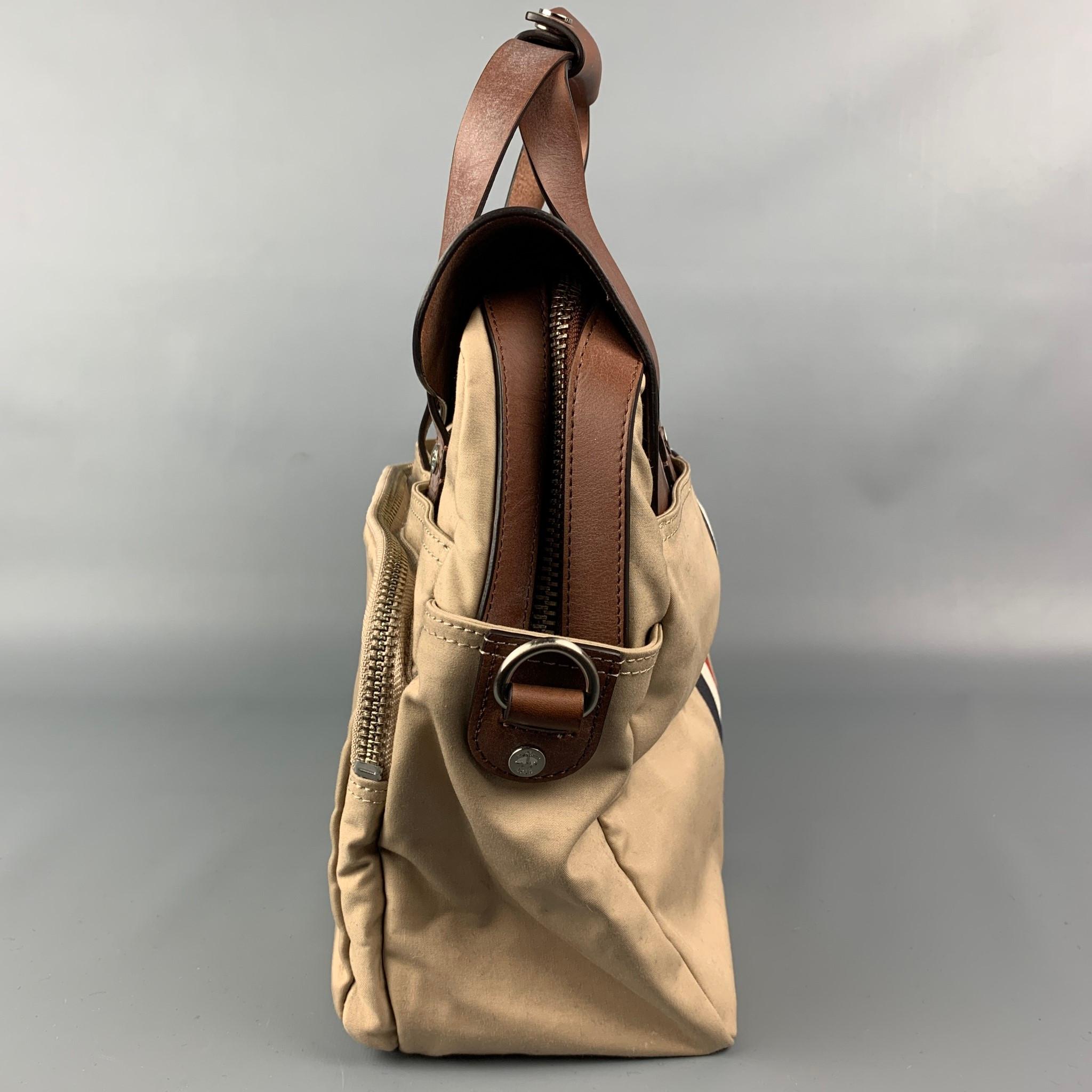 THOM BROWNE for BLACK FLEECE bag comes in a beige canvas with a tan leather trim featuring a messenger style, top handle, signature stripe detail, side pockets, silver tone hardware, zipper pocket, adjustable shoulder strap, snap button detail, and