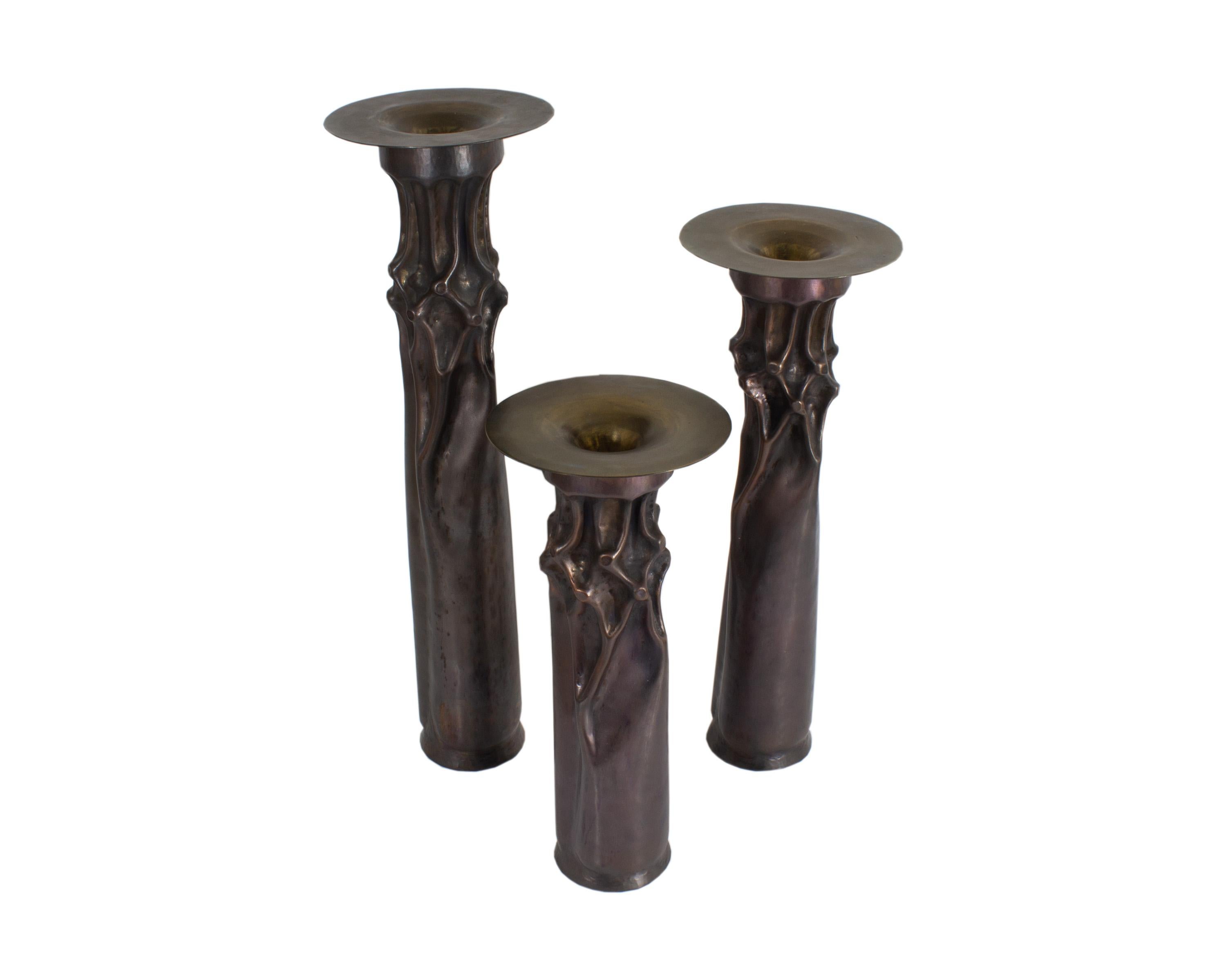 A set of three Brutalist candle holders designed by American artist Thomas Roy Markusen (born 1940). Composed of copper, each candle holder features a cylindrical shape with an abstracted sculptural design just below the flared holder. All are